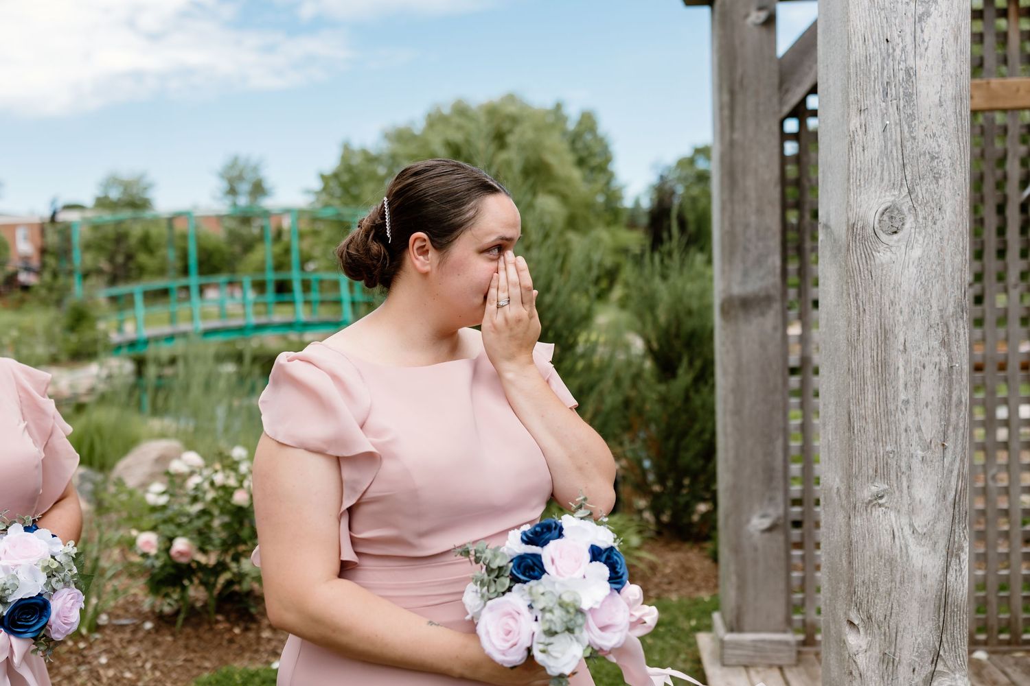 Emotional bridesmaid in pink dress wiping tears while holding floral bouquet during outdoor wedding ceremony.