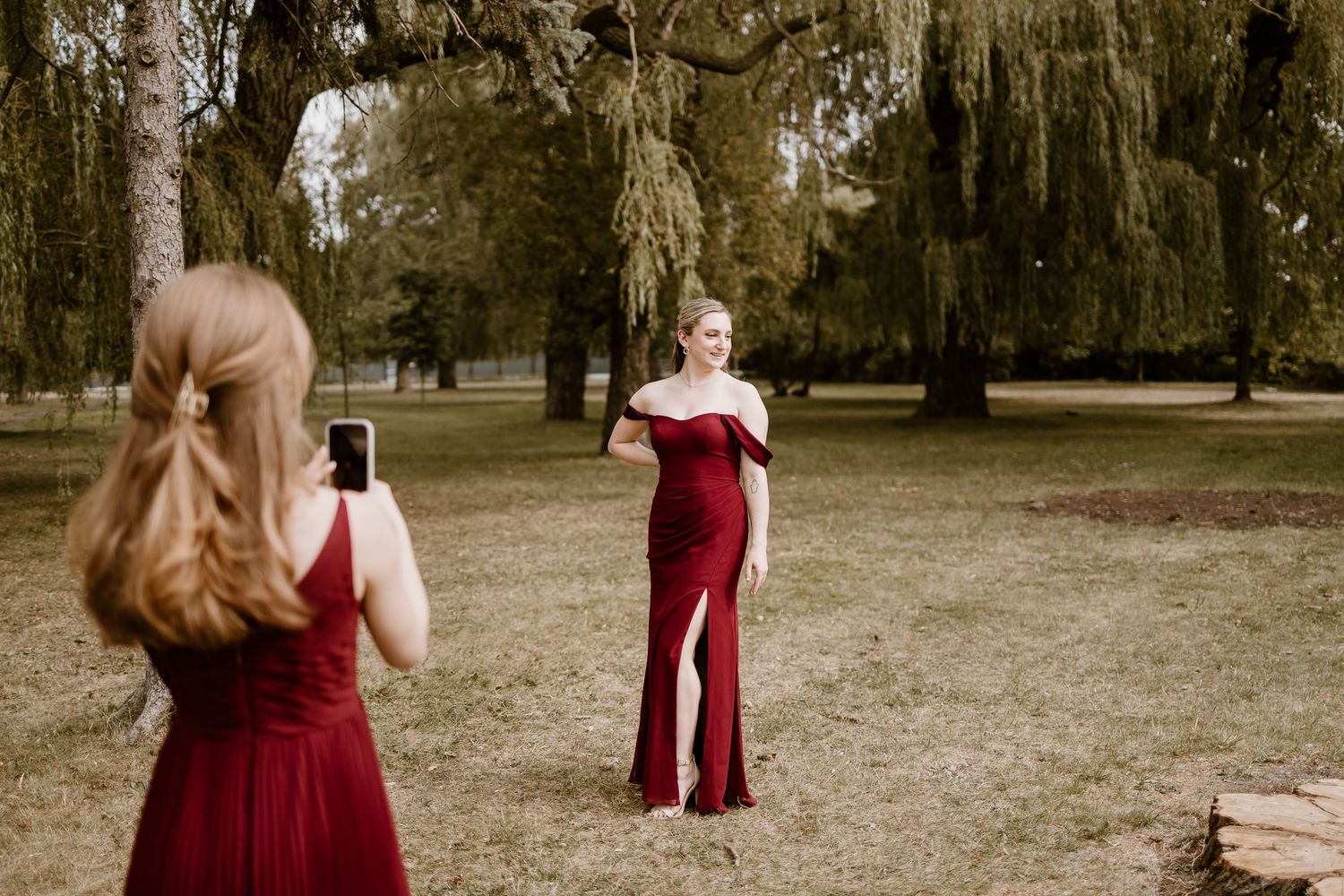 Two people in burgundy dresses taking photos in a park with moss-covered trees.