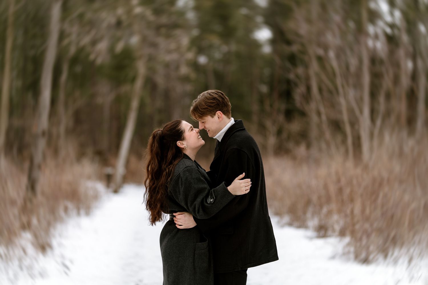 A romantic winter photoshoot showing a couple in black coats embracing in the snow.