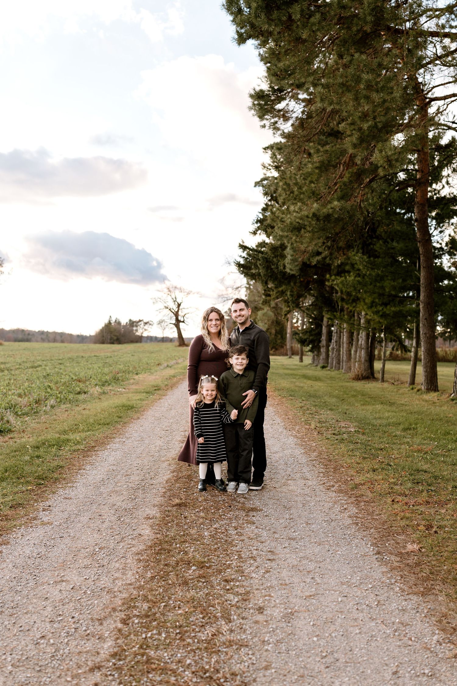Family walking together on a gravel path lined with pine trees at sunset.