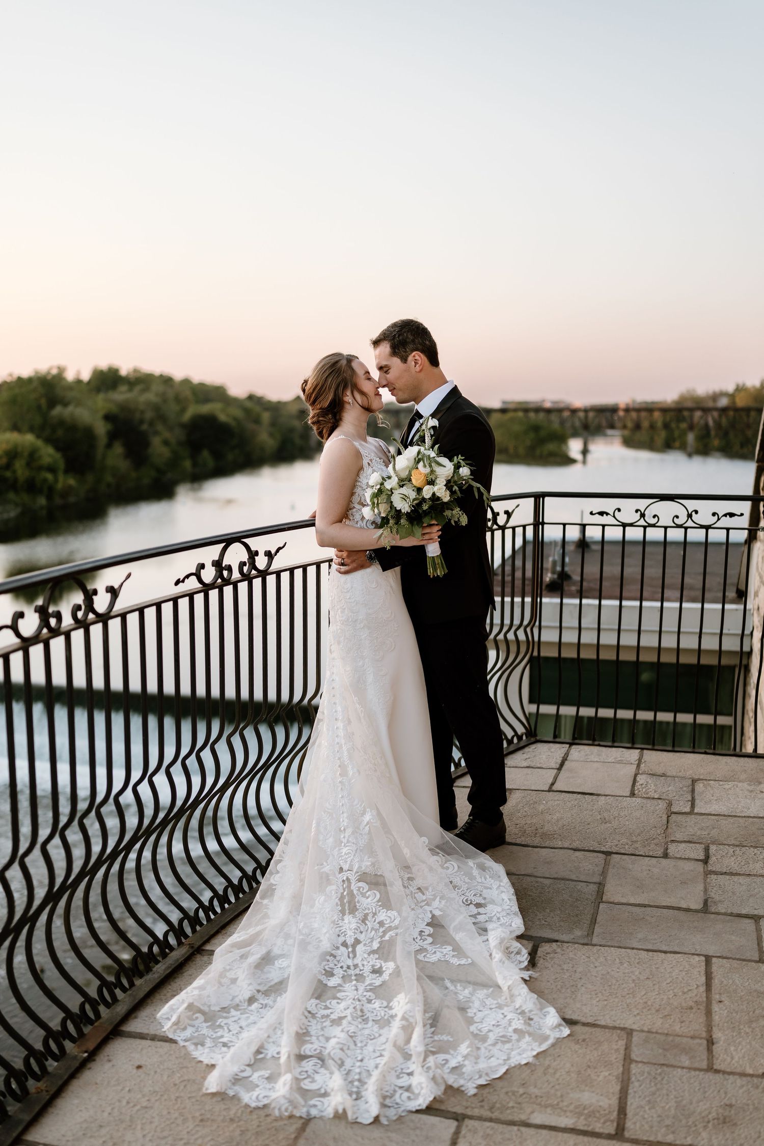 Newlyweds posing by ornate iron railings overlooking a scenic waterfront view.