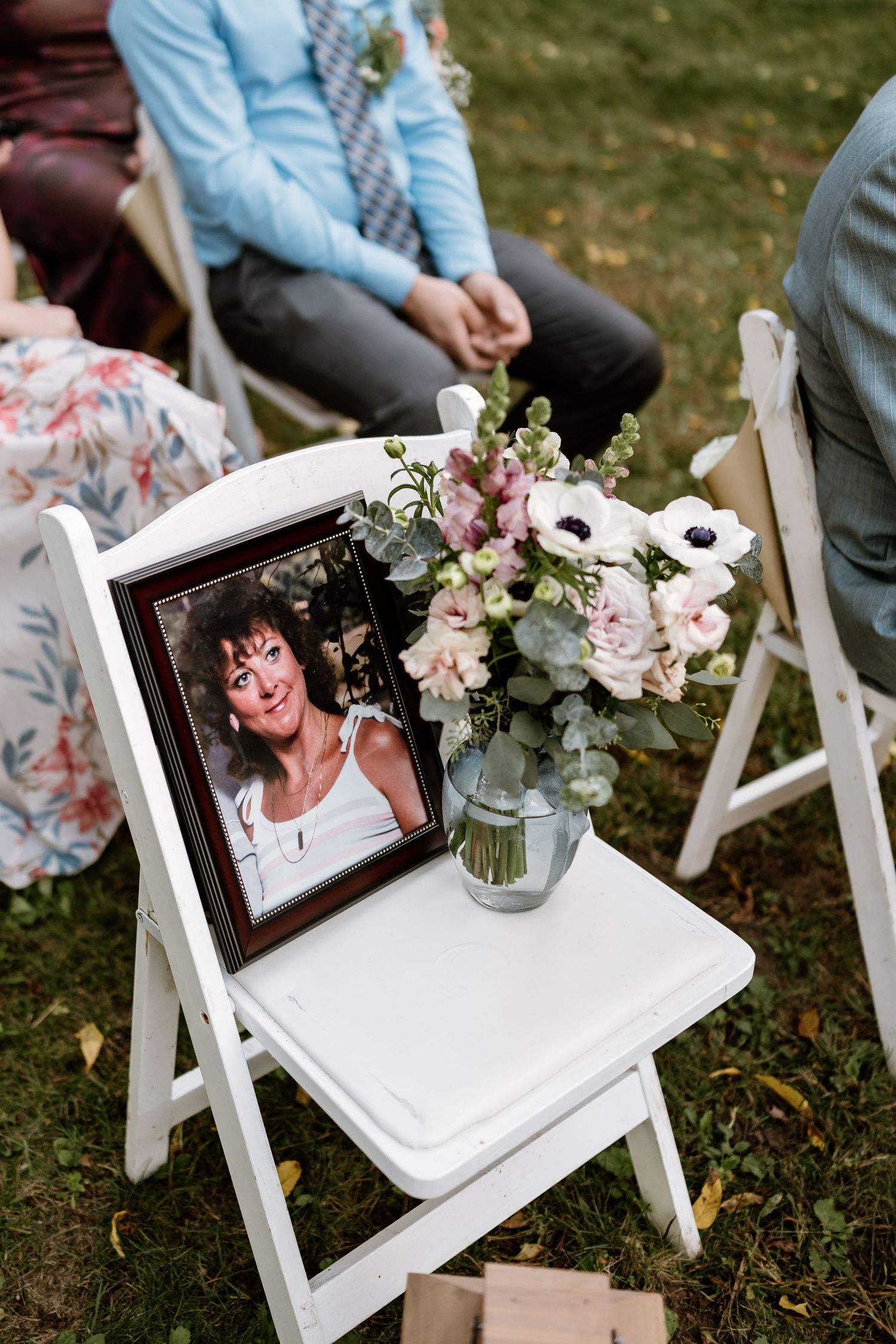 Memorial photo display on white folding chair with pink floral arrangement at outdoor wedding ceremony.