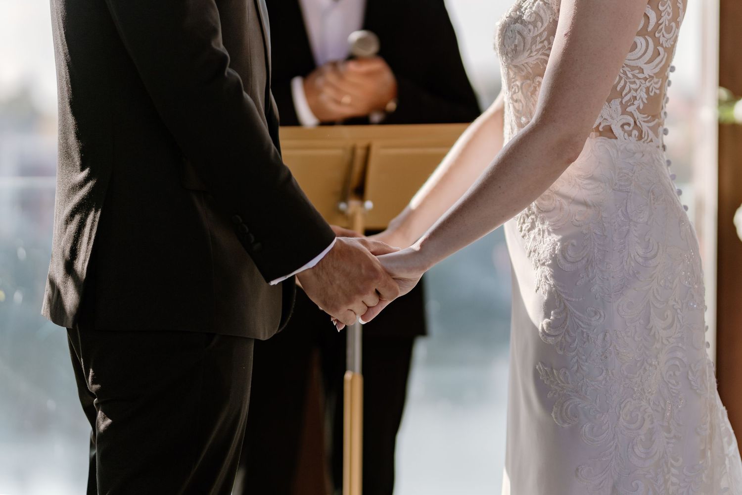 Close-up of couple holding hands during wedding ceremony with lace dress details.
