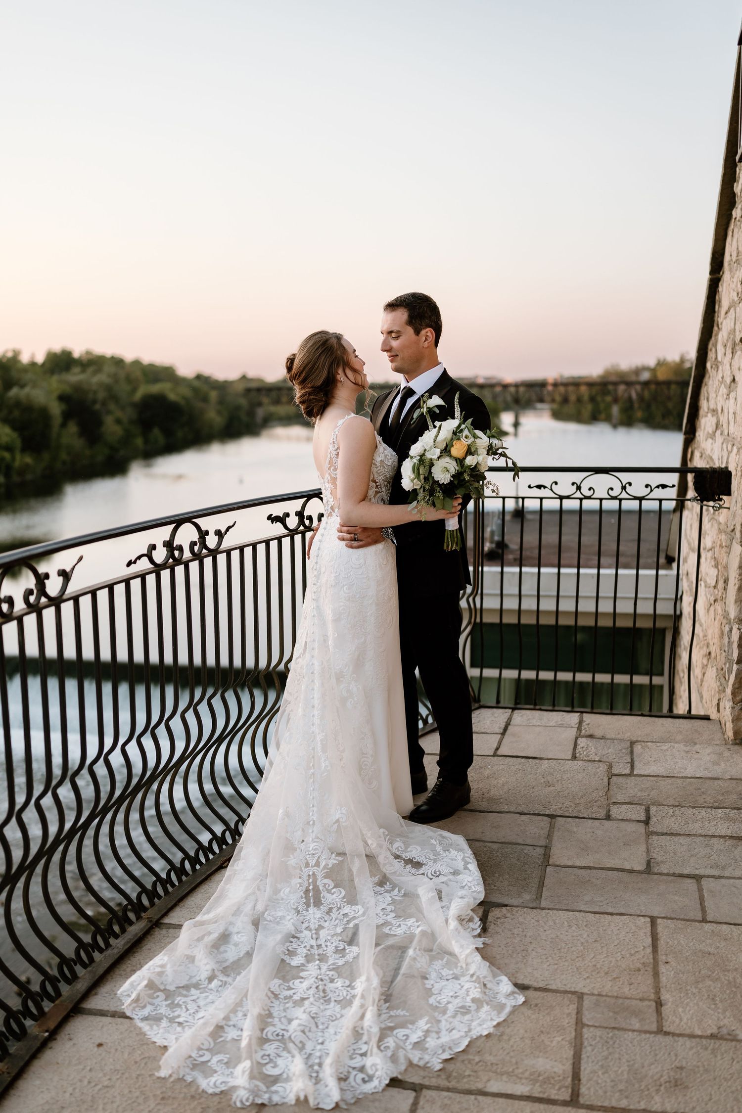 Elegant wedding couple poses on stone balcony overlooking waterfront at sunset.