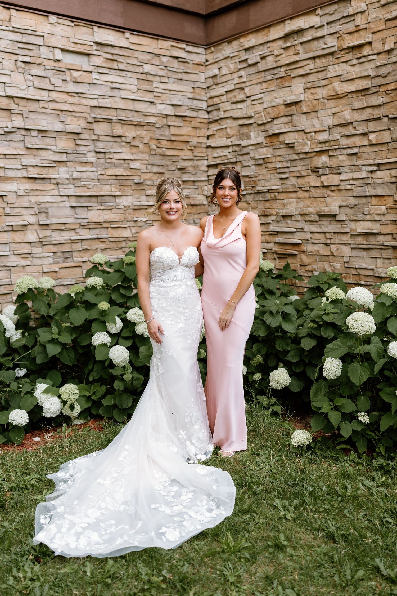 A bride in a floral lace gown poses with a bridesmaid in a pink dress near white hydrangeas.