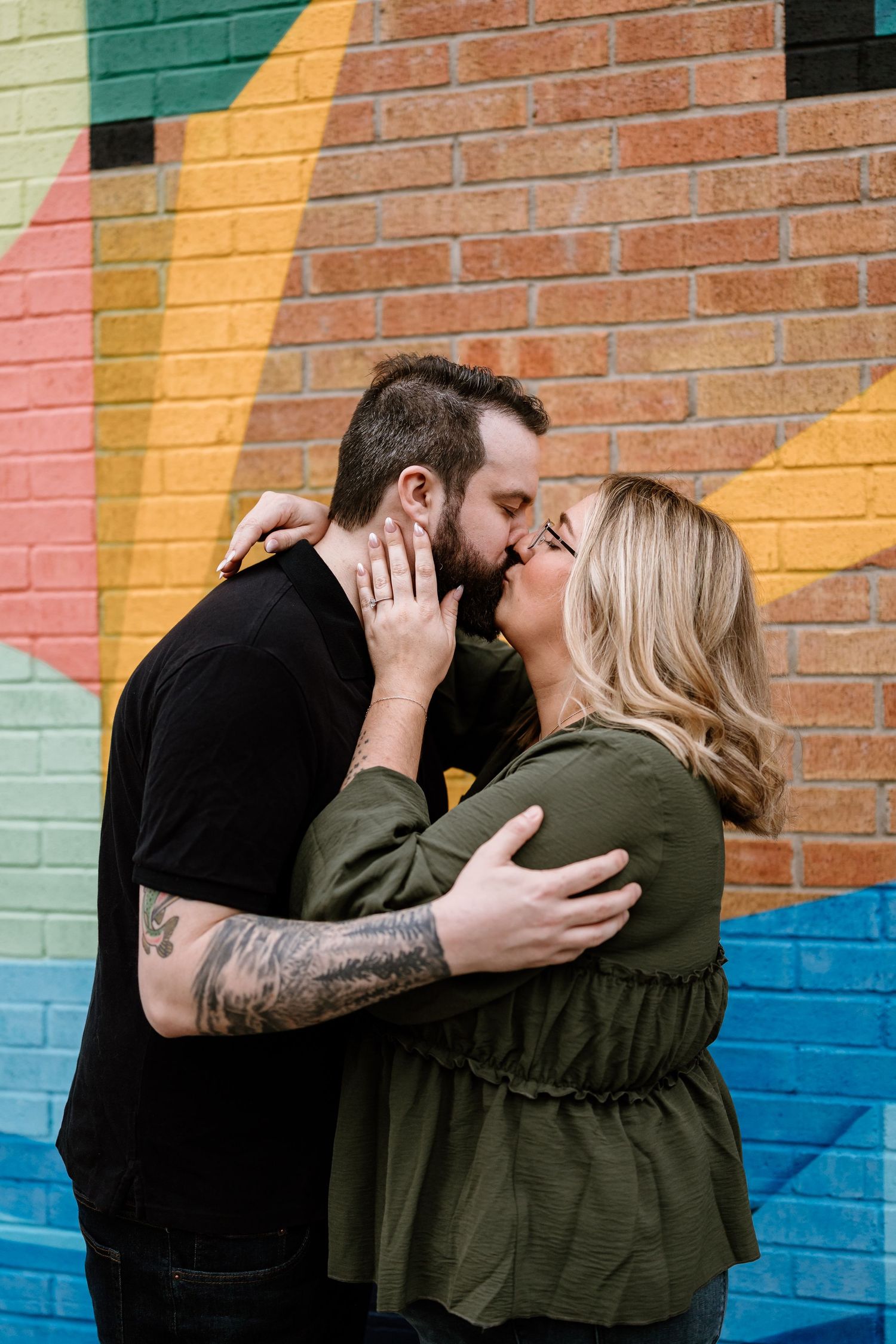 A couple shares a romantic kiss in front of a colorful brick mural, the man with tattoos embracing a blonde woman.