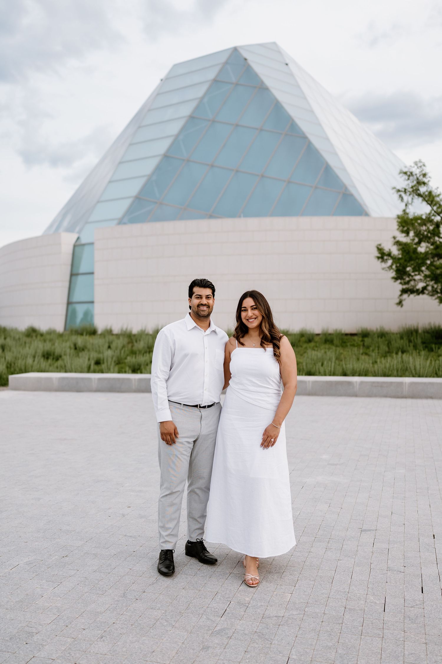 Couple in white attire posing in front of a modern geometric glass building on a cloudy day.