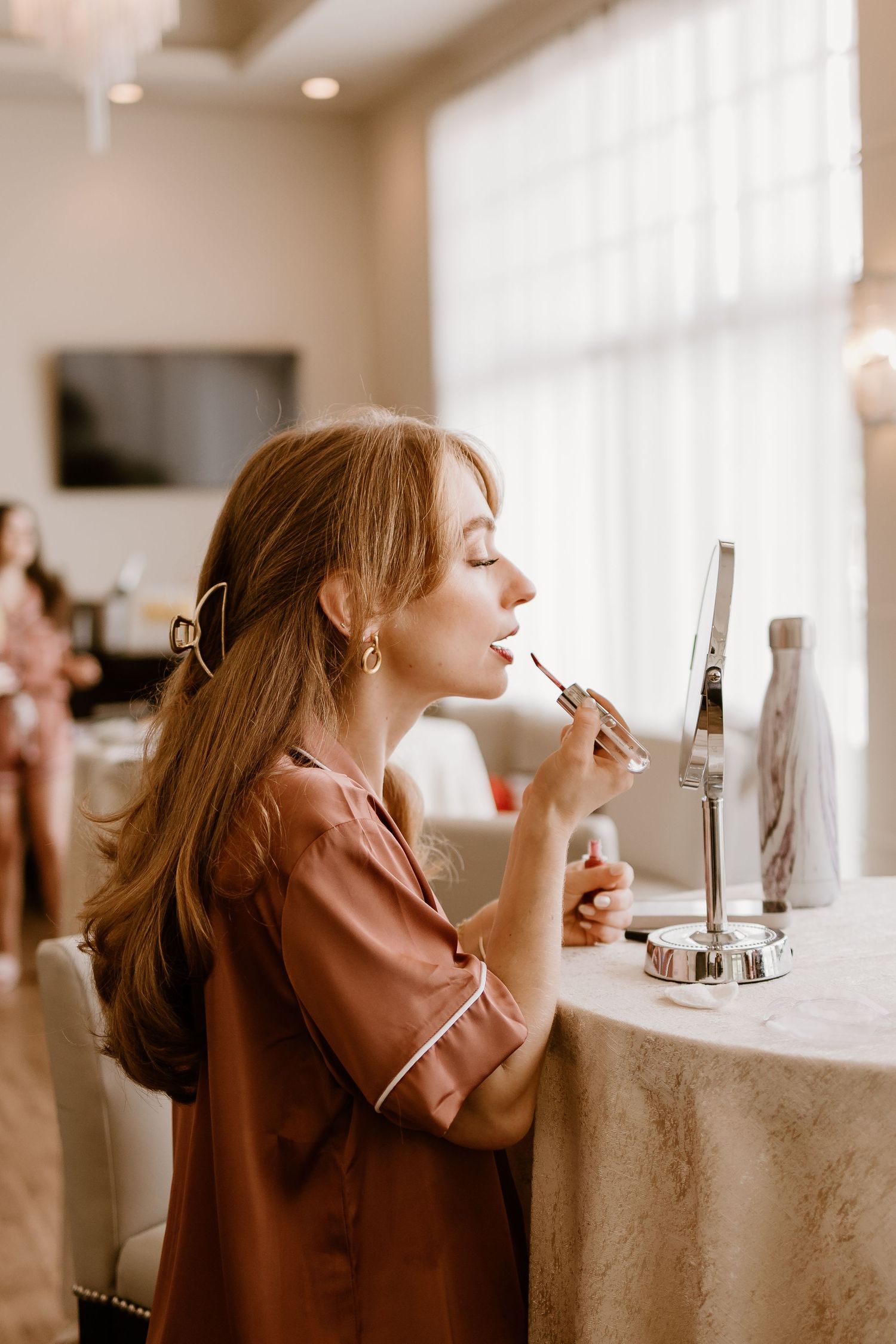 Side profile of woman applying lipstick at vanity table.