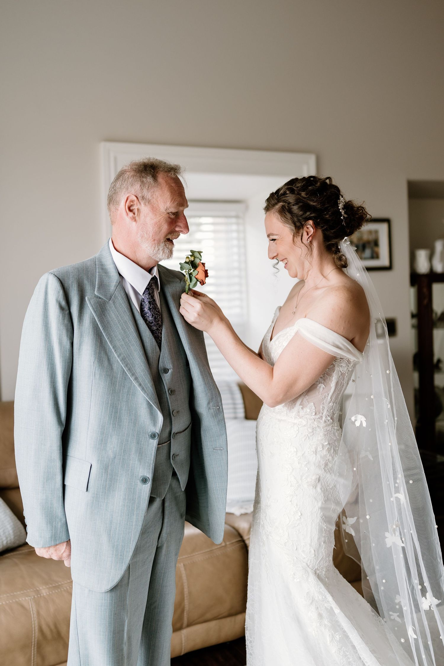 A tender moment between a bride in white and someone wearing a light gray suit adjusting a boutonniere.