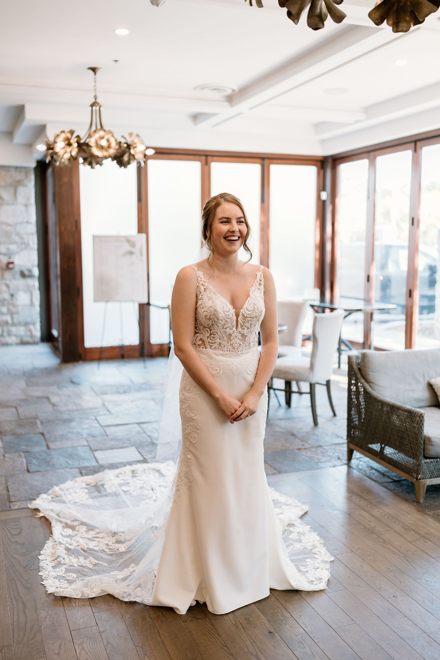 A bride wearing a beautiful lace wedding gown stands in an elegant room with large windows.