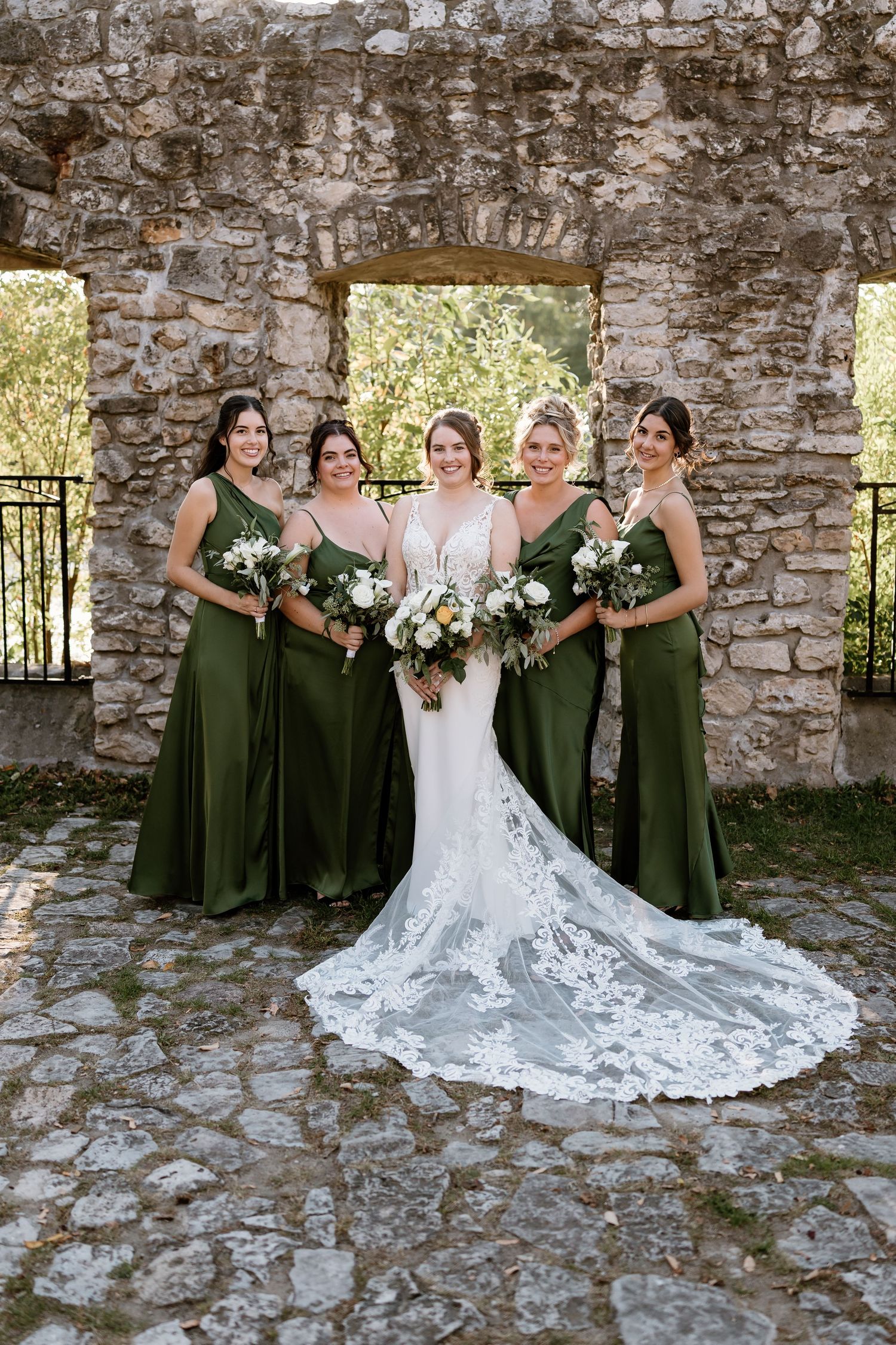Bride in lace gown poses with bridesmaids wearing olive green dresses against stone wall.