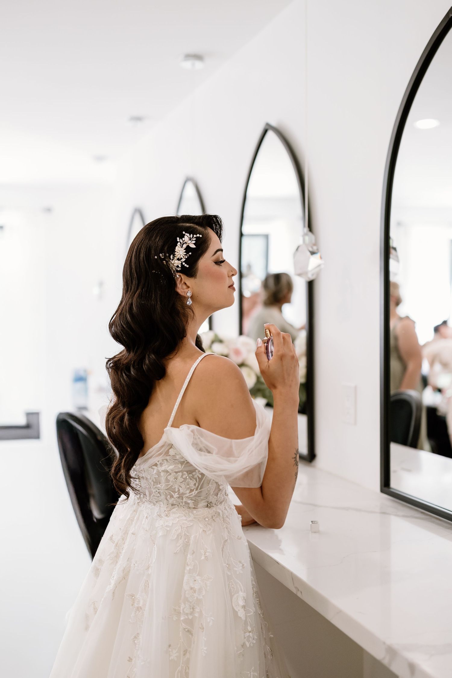 Bride in a lace gown applying perfume at a vanity mirror with floral hair accessories before wedding.