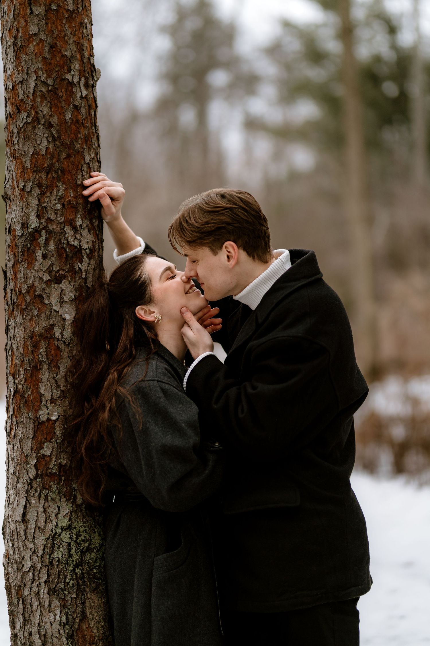 A couple shares a romantic moment against a pine tree in a snow-covered forest.
