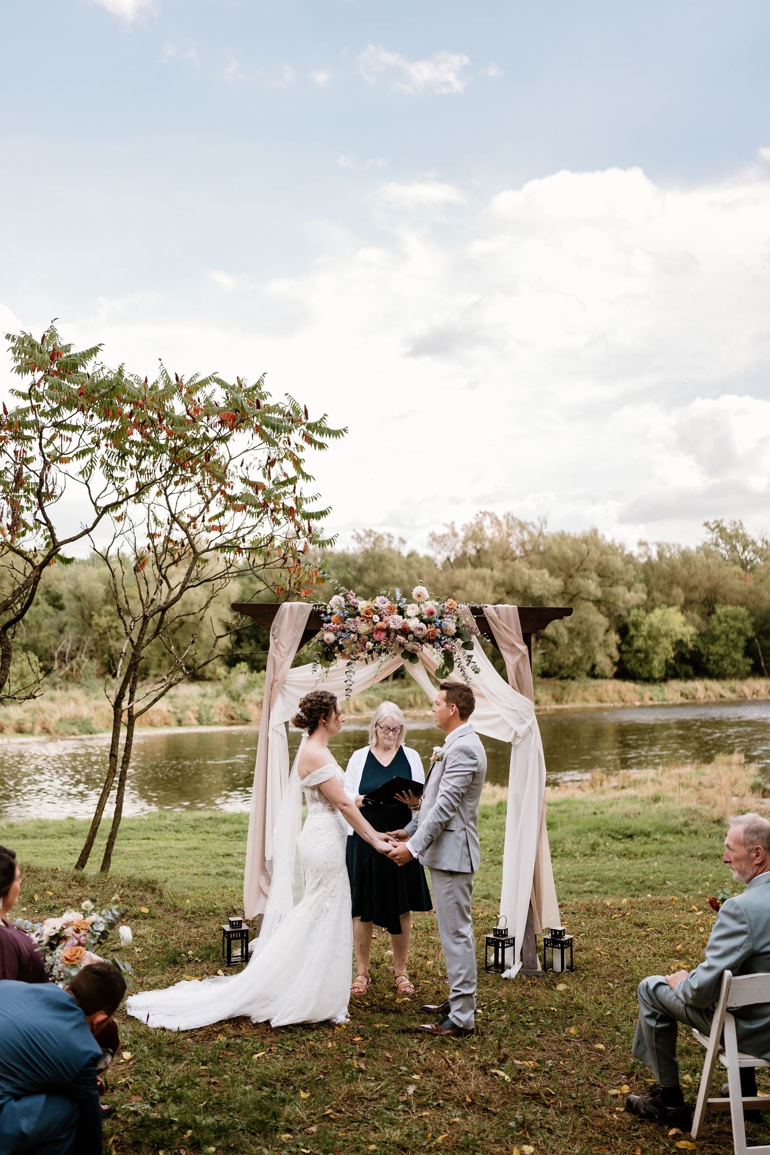 Outdoor wedding ceremony by lake with white arch, floral arrangements and folding chairs.