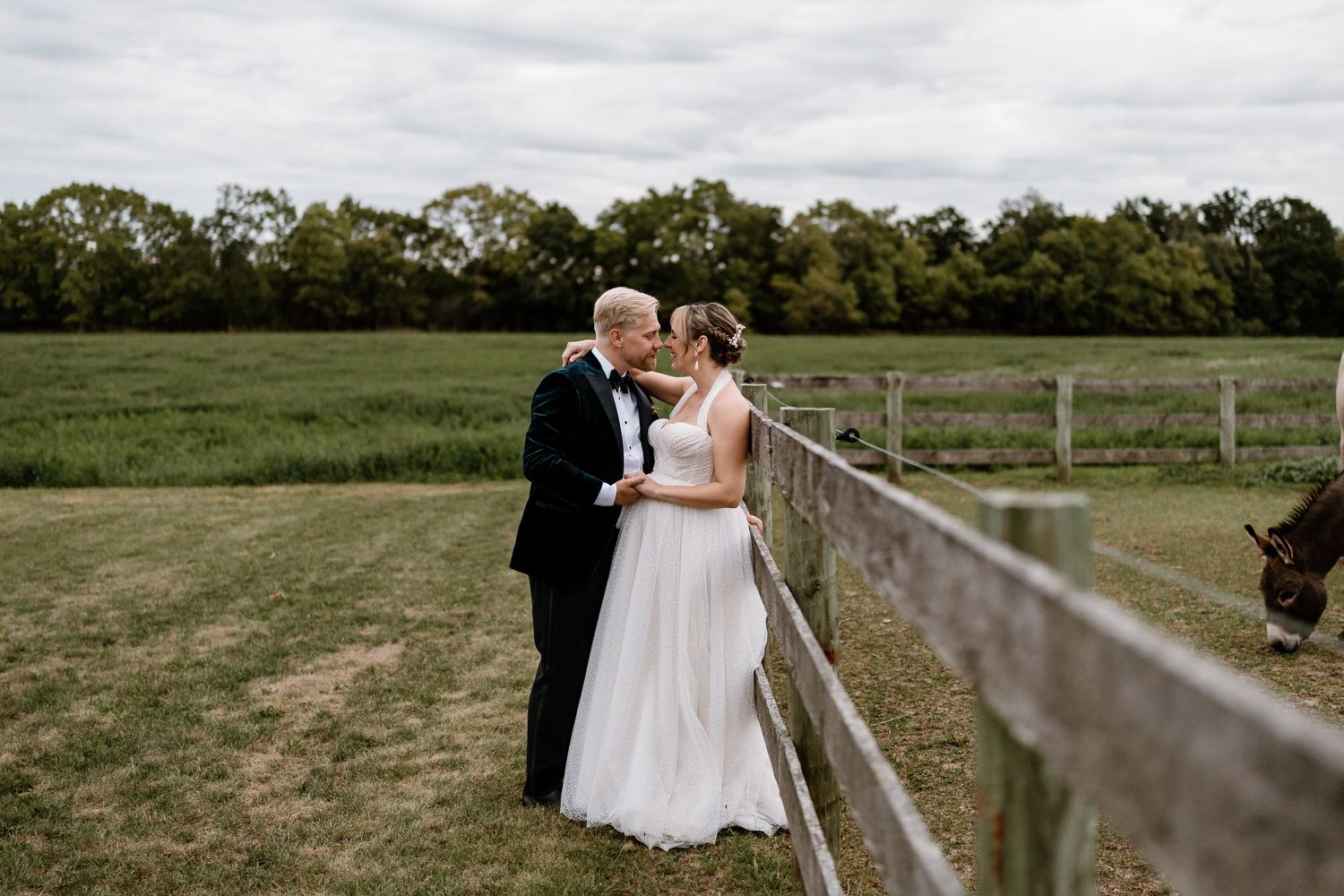 Newlyweds share a tender moment by a wooden fence in a pastoral field.