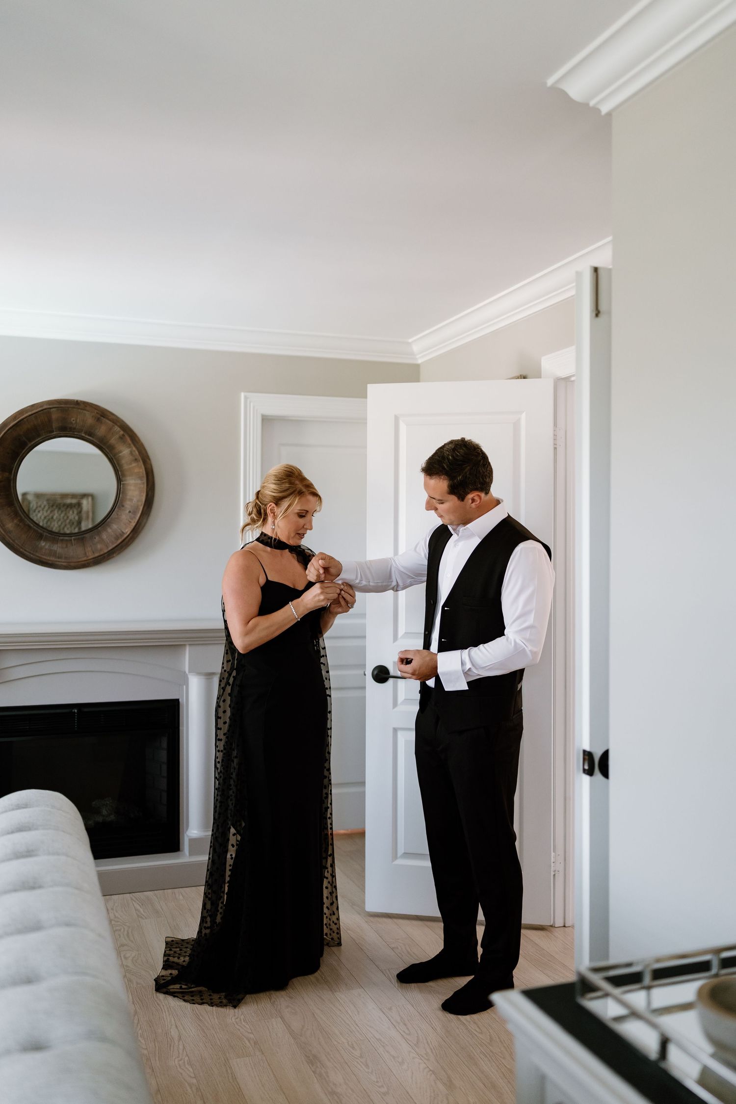 Two people in formal attire getting ready in an elegant room with white walls and round mirror.