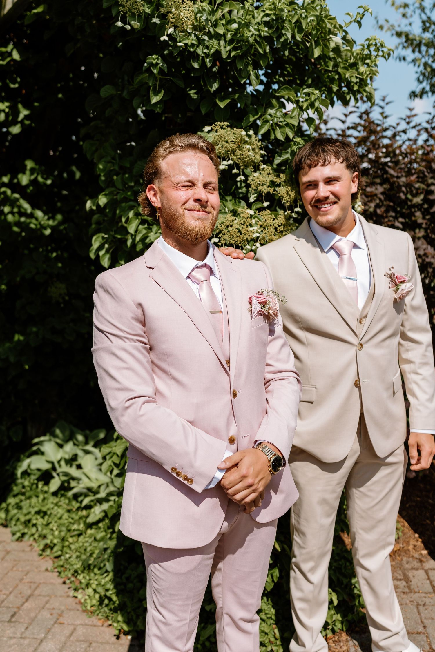 Two groomsmen in pink and beige suits smiling outdoors on sunny wedding day.
