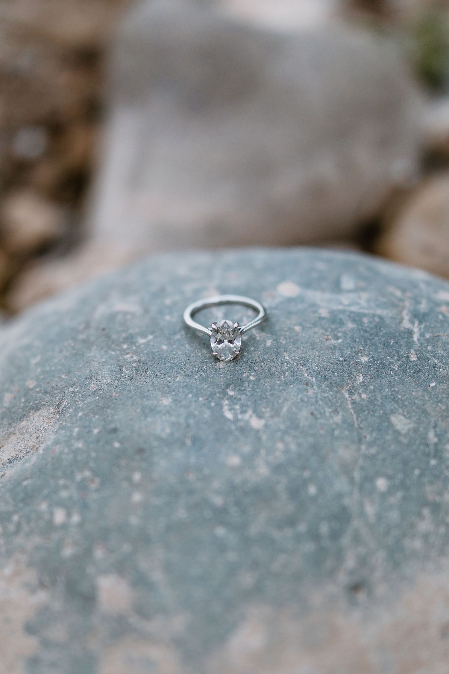 Oval diamond solitaire engagement ring displayed on a smooth gray rock outdoors.