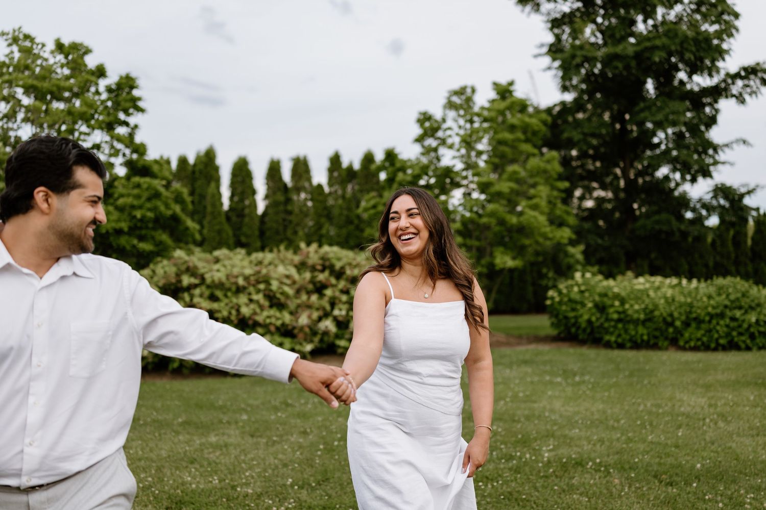 A joyful couple in white attire holds hands while walking through a lush green garden, smiling at each other.