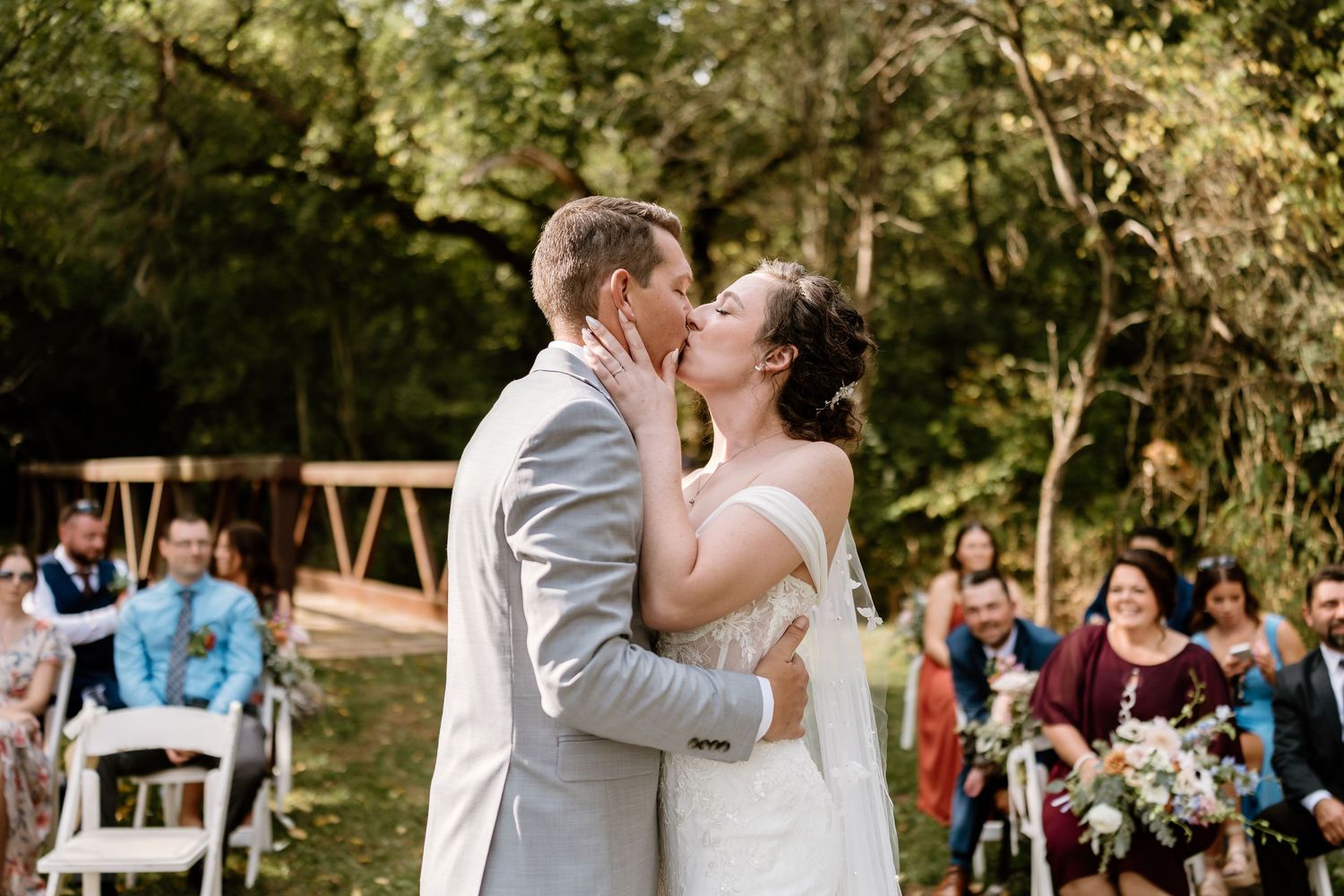 Outdoor wedding ceremony in wooded setting with guests watching couple exchange vows.