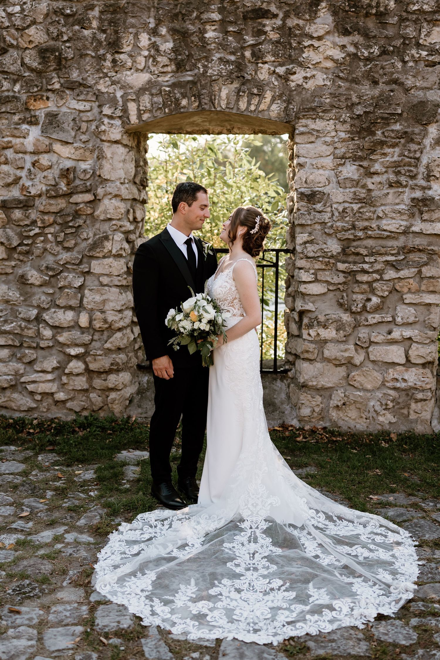 Bride's dramatic lace train spreads across stone courtyard during couple's portrait session.