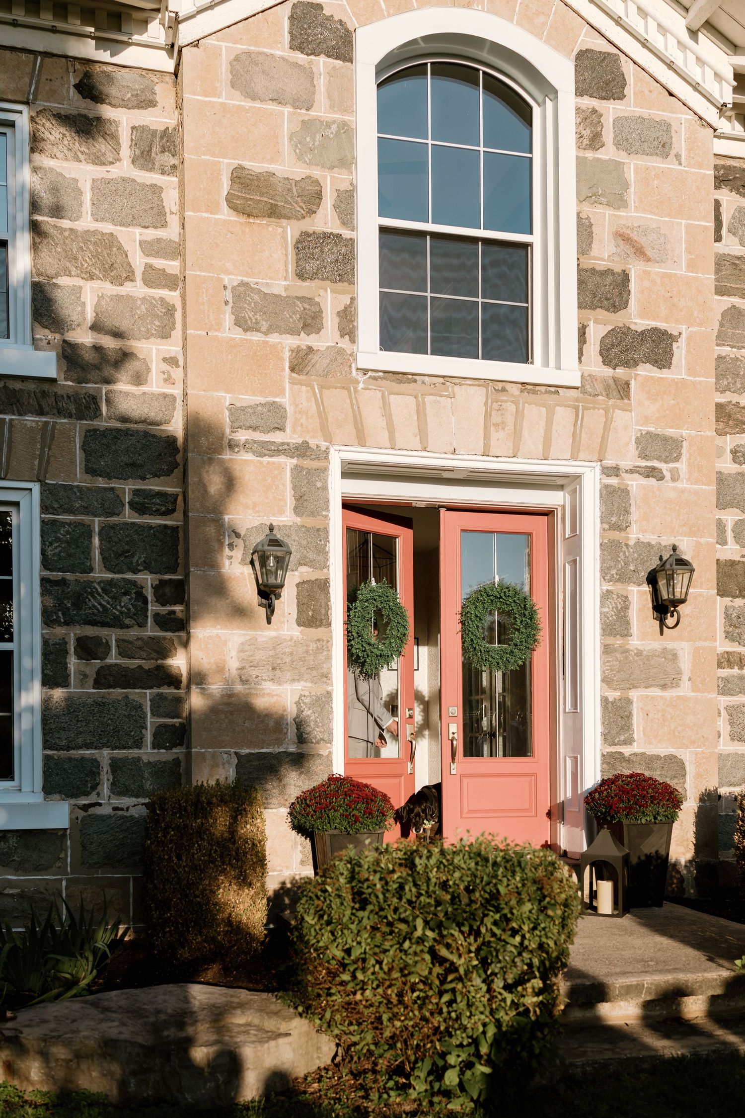 A stone building exterior with pink doors, white windows, and decorative bushes and wreaths.