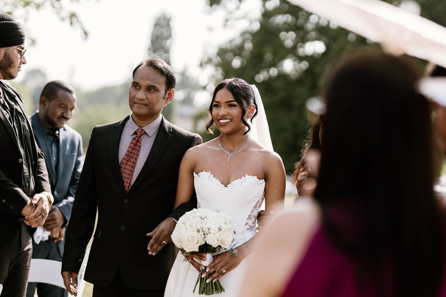 A bride in a white strapless gown holds a bouquet during an outdoor wedding ceremony.
