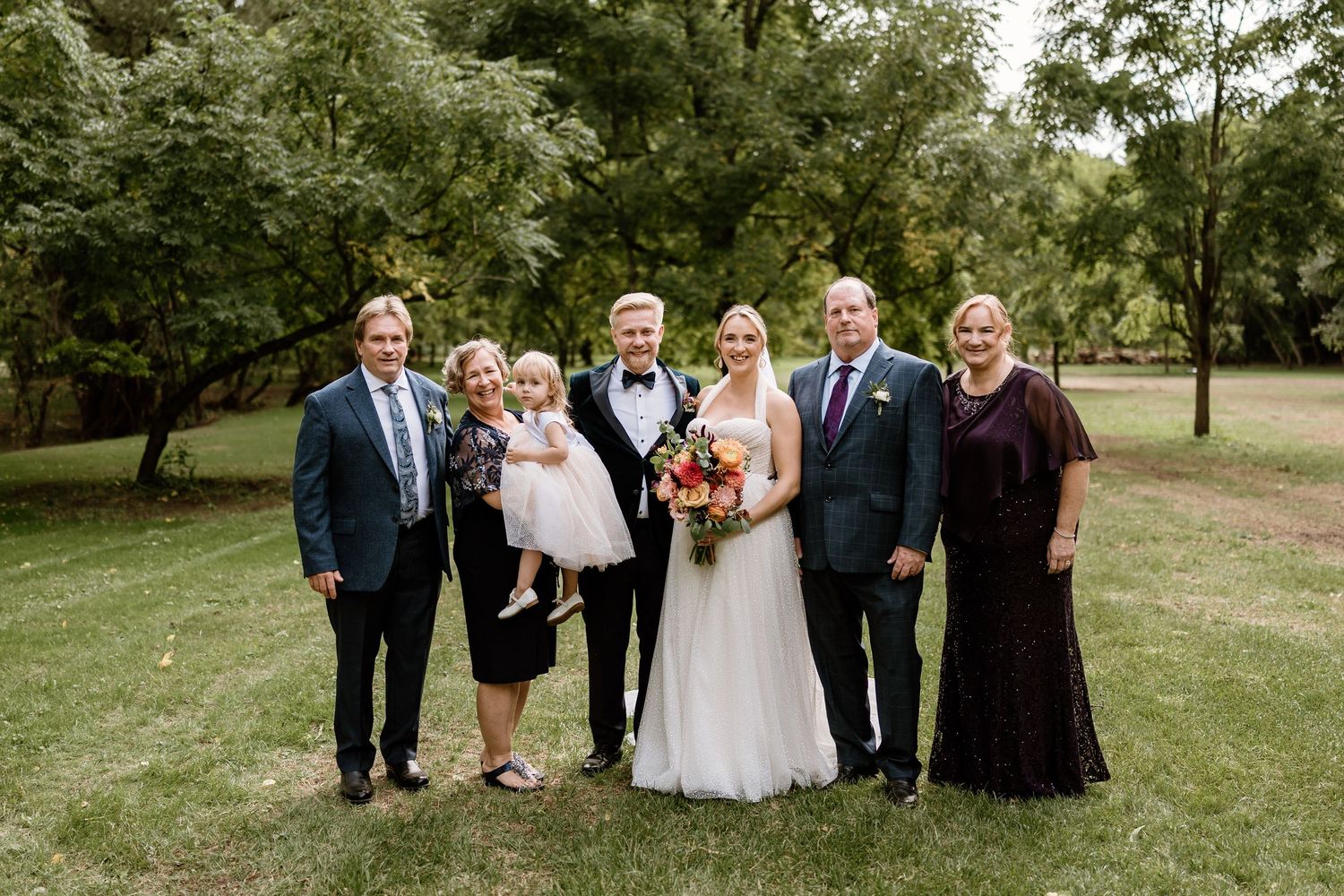 Wedding party poses for formal group photos on grassy lawn with trees in background.