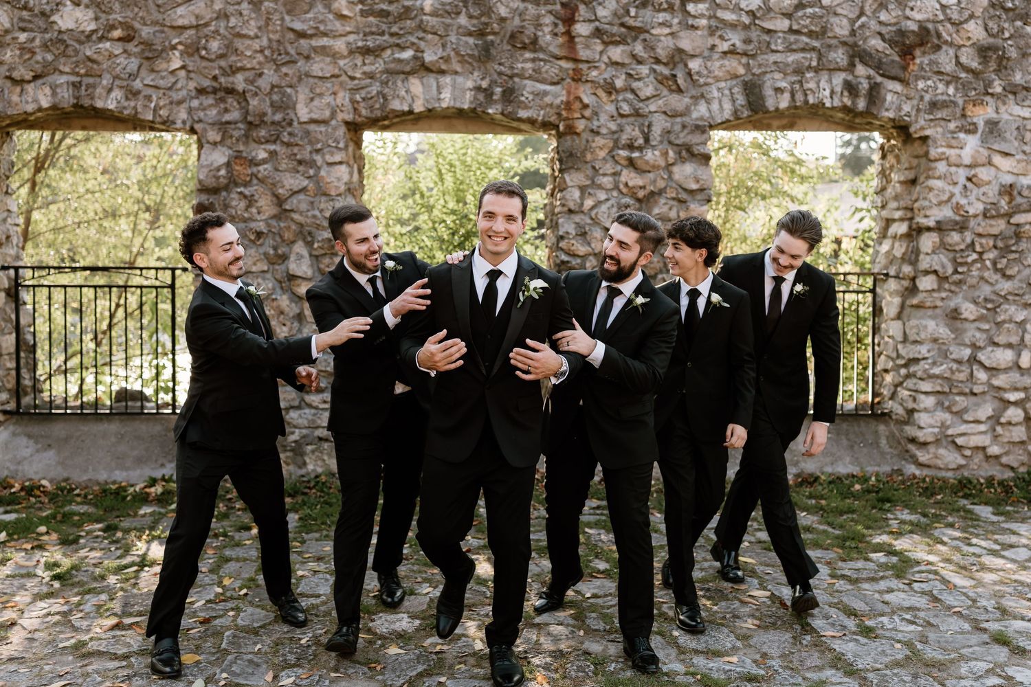 Groomsmen in black suits share candid laughs against stone wall backdrop.