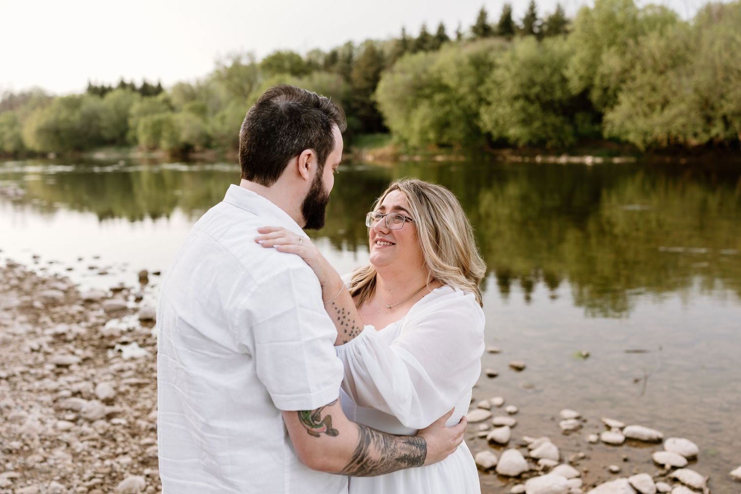 Couple in white outfits embracing and smiling by a rocky riverbank with lush green trees in the background.
