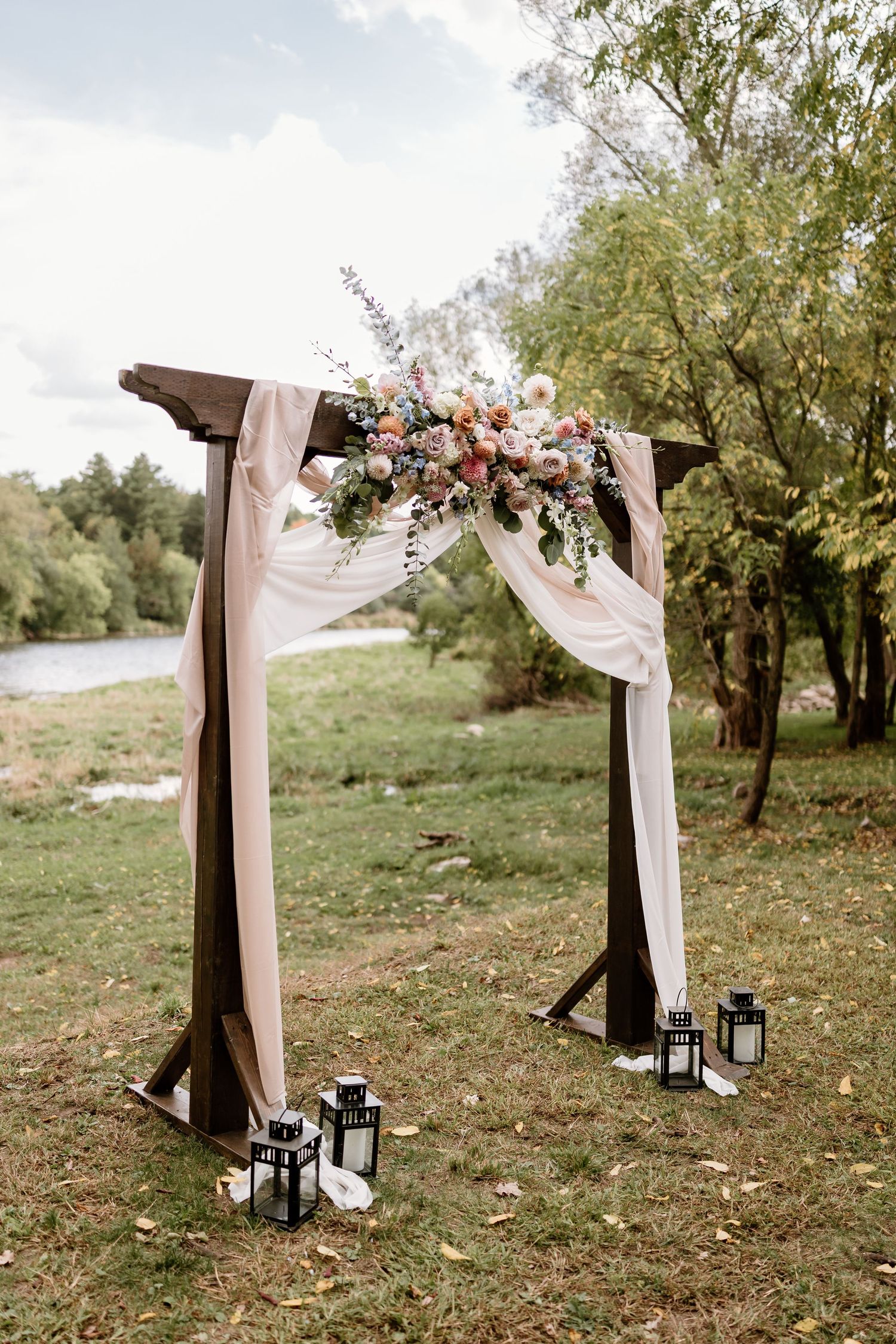 A wooden wedding arch decorated with pink flowers, white fabric draping, and lanterns in an outdoor setting.