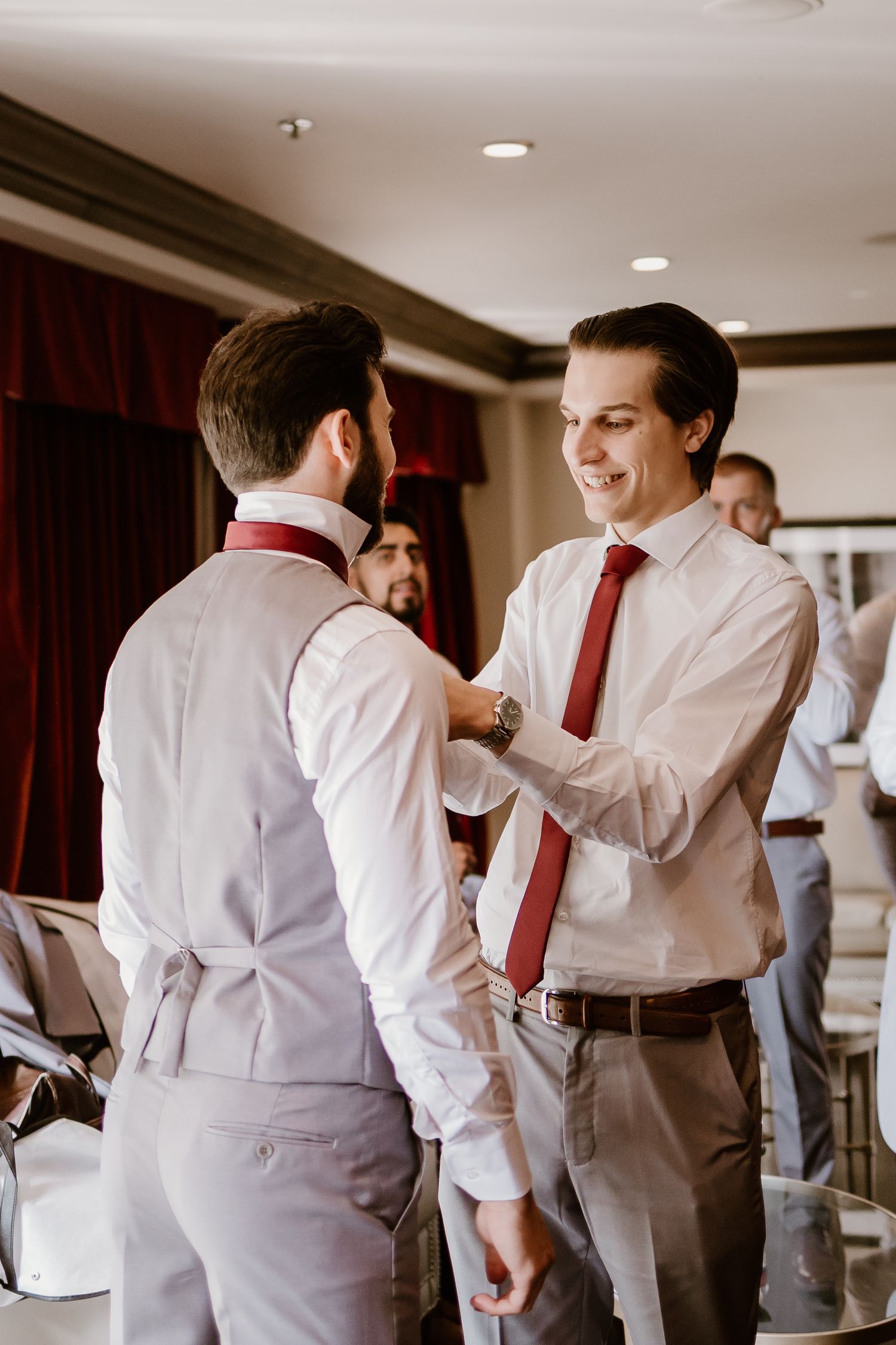 Groomsmen in white shirts and vests help each other prepare for wedding ceremony.