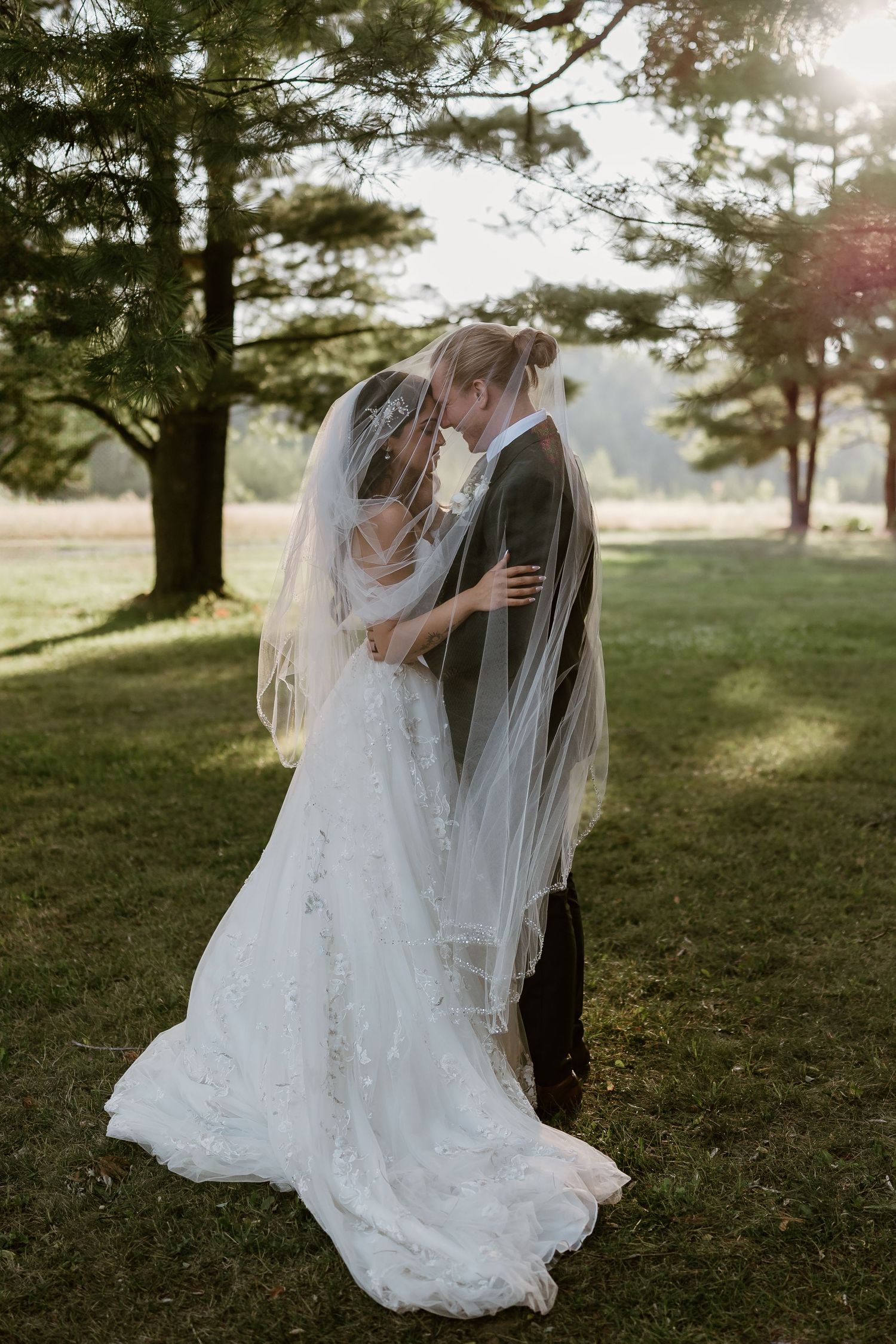 Wedding couple embracing on grassy field with pine trees at sunset.