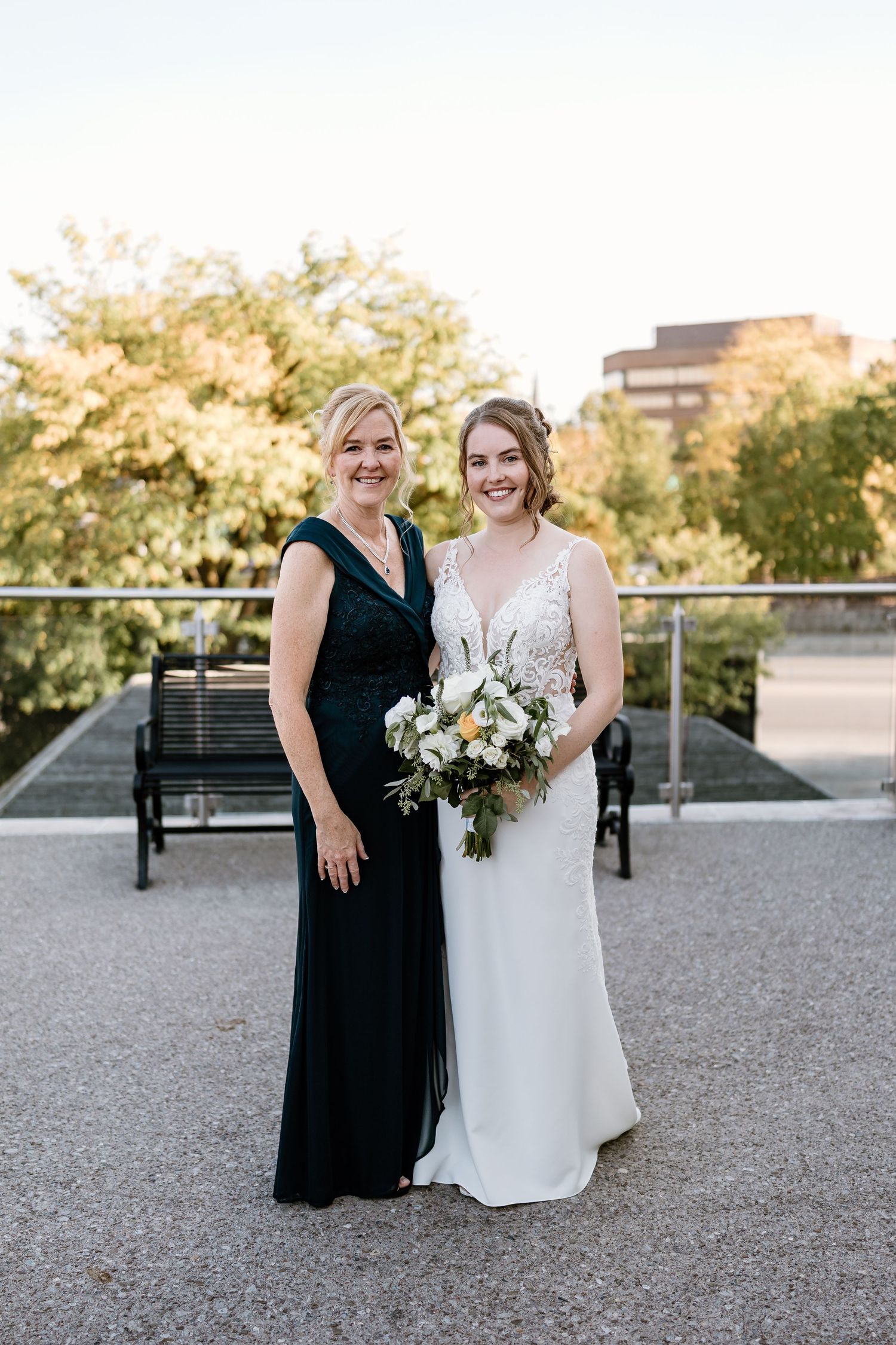 Two people in formal attire pose with bouquet on modern outdoor terrace.