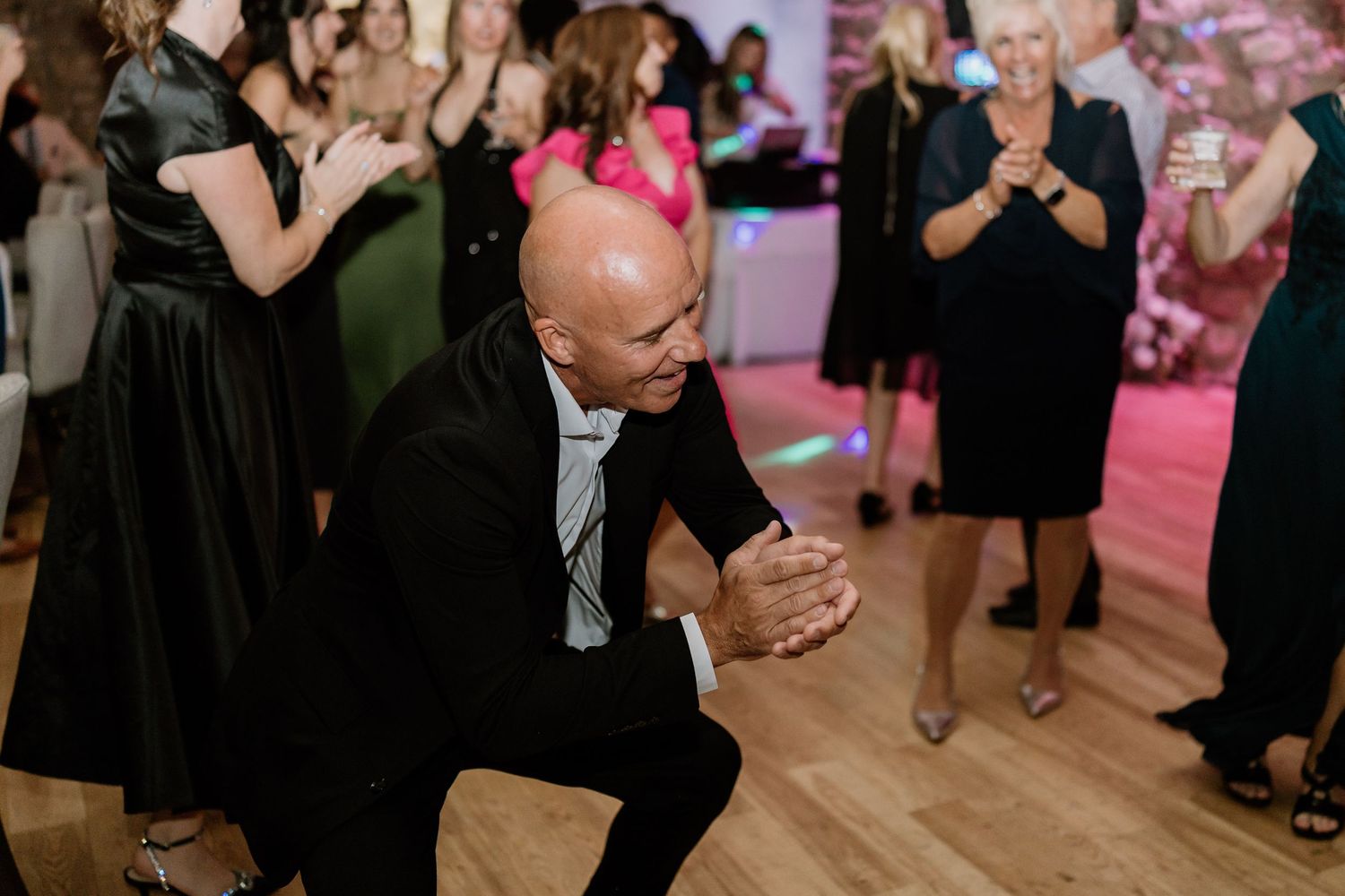 Lively dance floor scene at wedding reception with pink uplighting and stone backdrop.