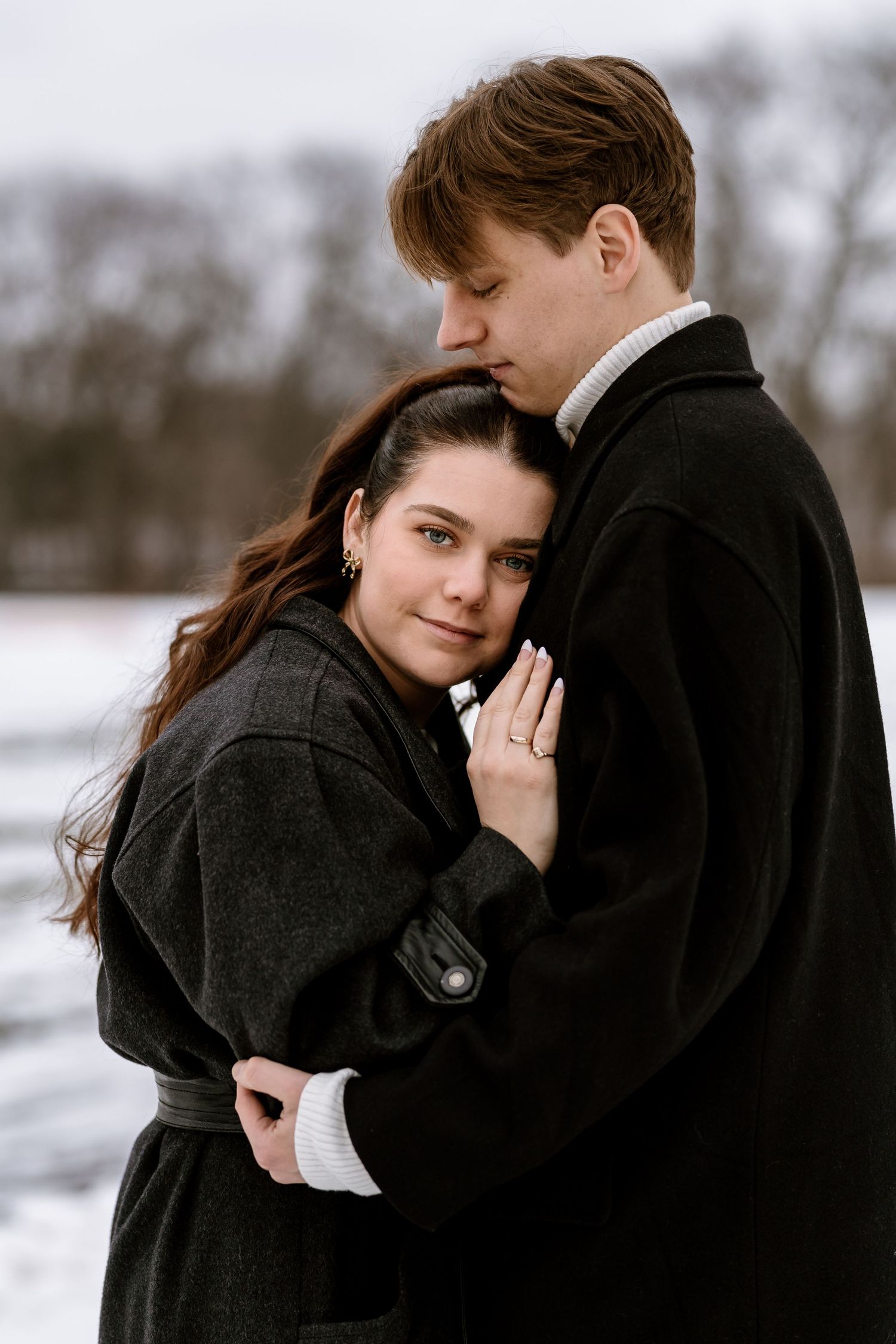 Romantic winter engagement photo of couple embracing in black coats against snowy backdrop.