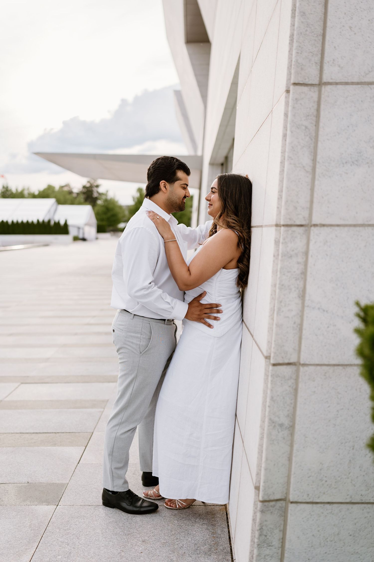 A couple in white attire shares a romantic moment leaning against a marble wall outdoors.
