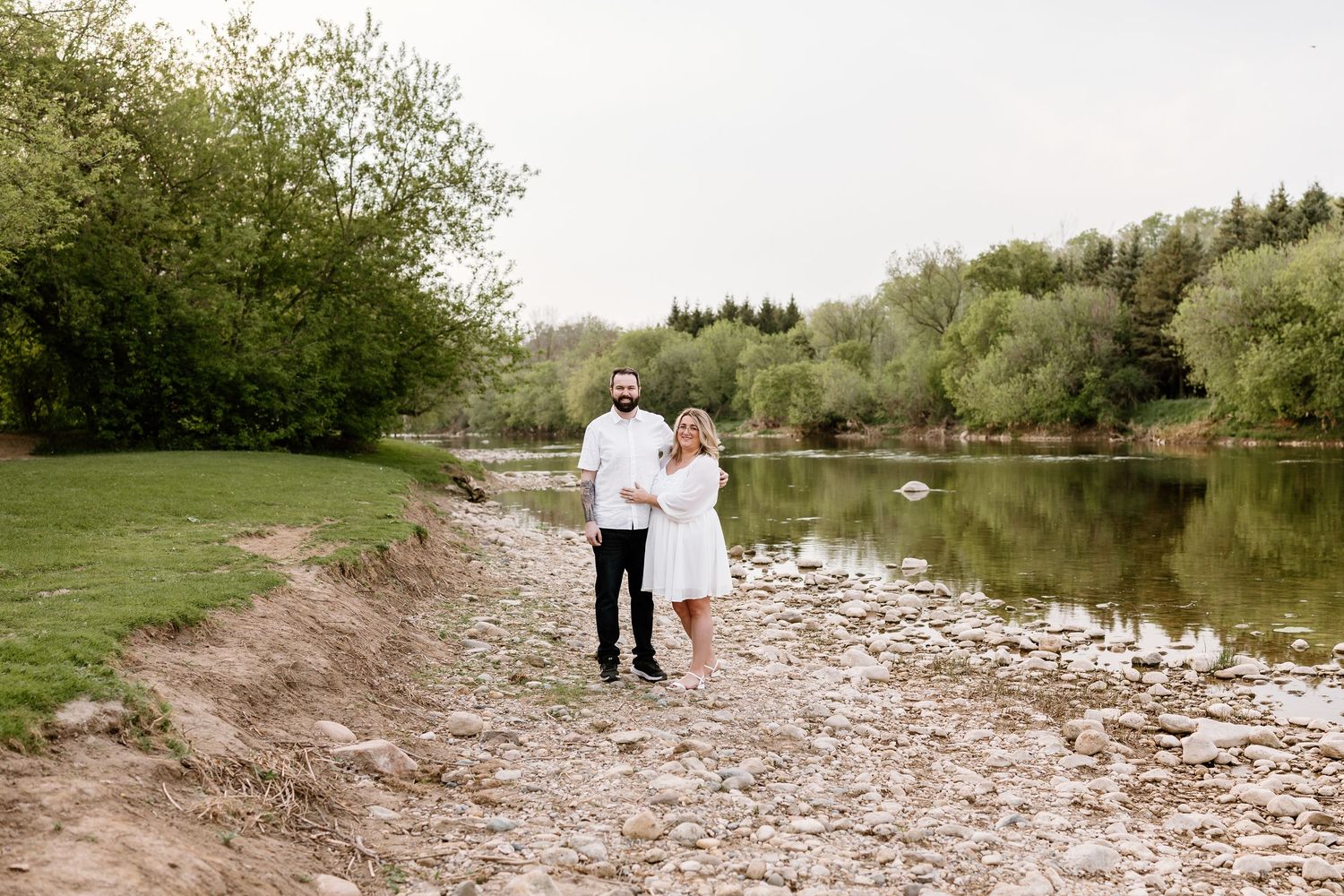 Couple holding hands on rocky riverbank surrounded by lush green trees during an outdoor engagement photo session.
