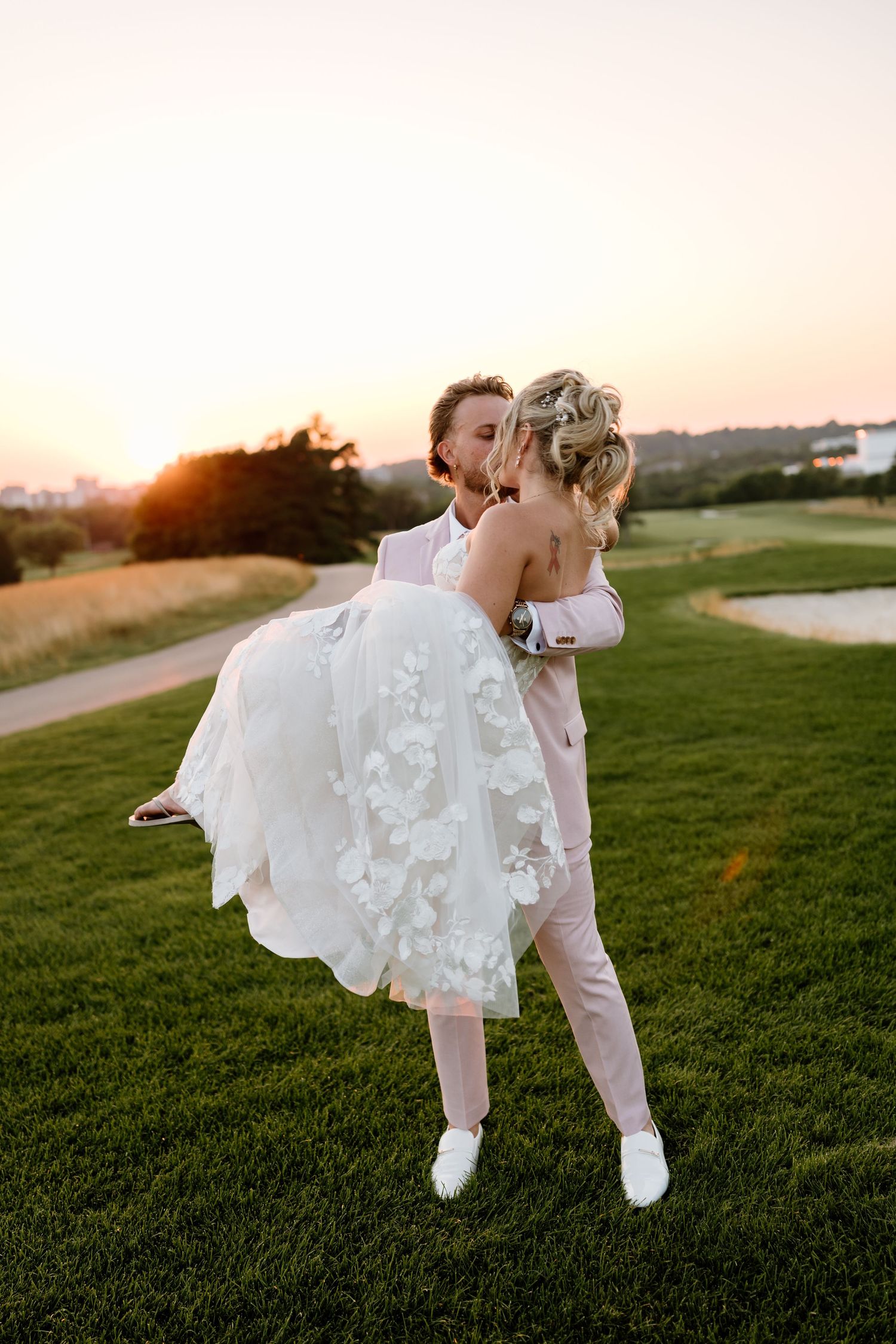 Groom in pink suit lifts bride in floral white dress at sunset on lush green golf course.