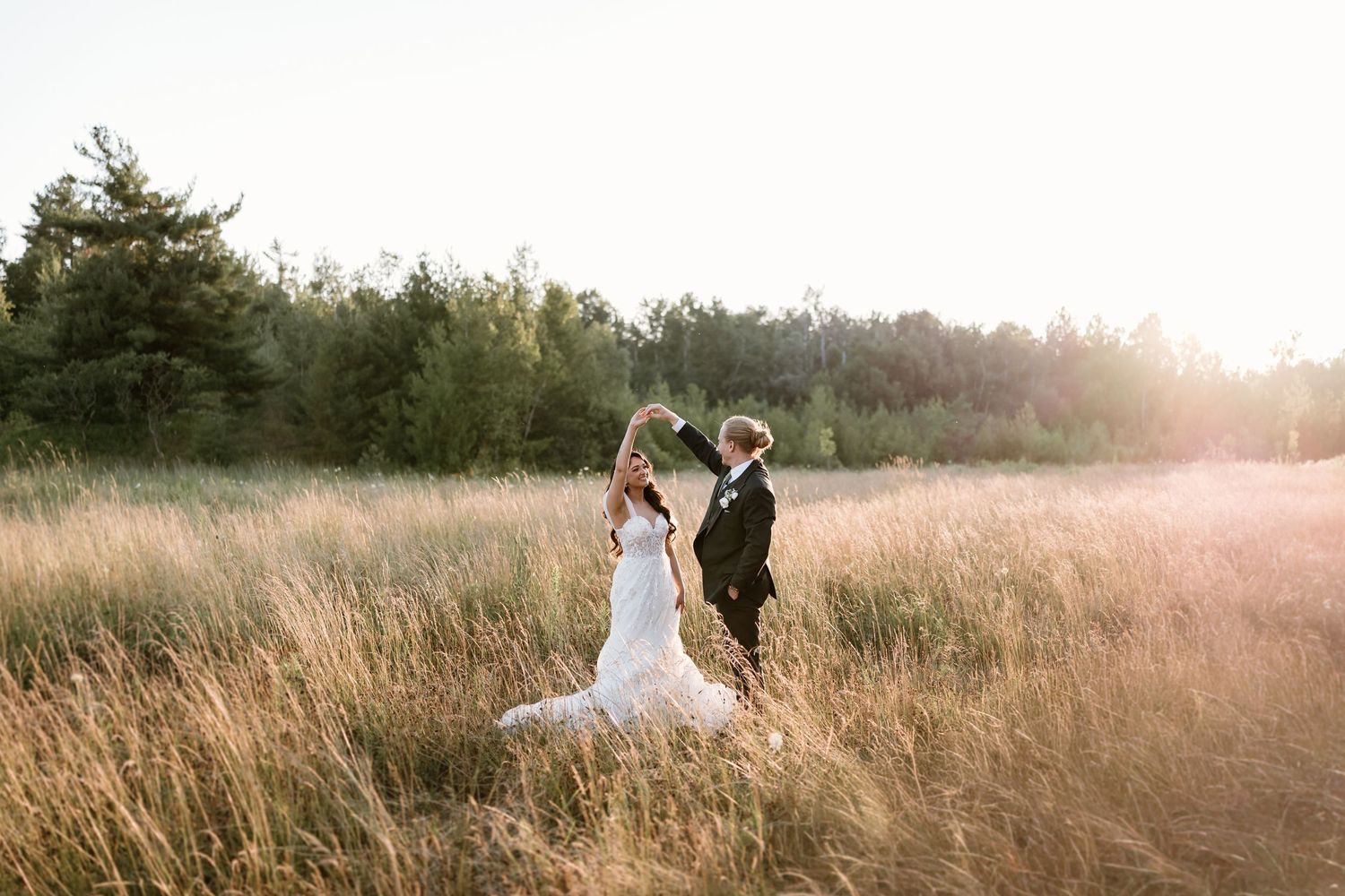 Bride and groom dancing joyfully in a golden field at sunset, surrounded by tall grass and lush trees.