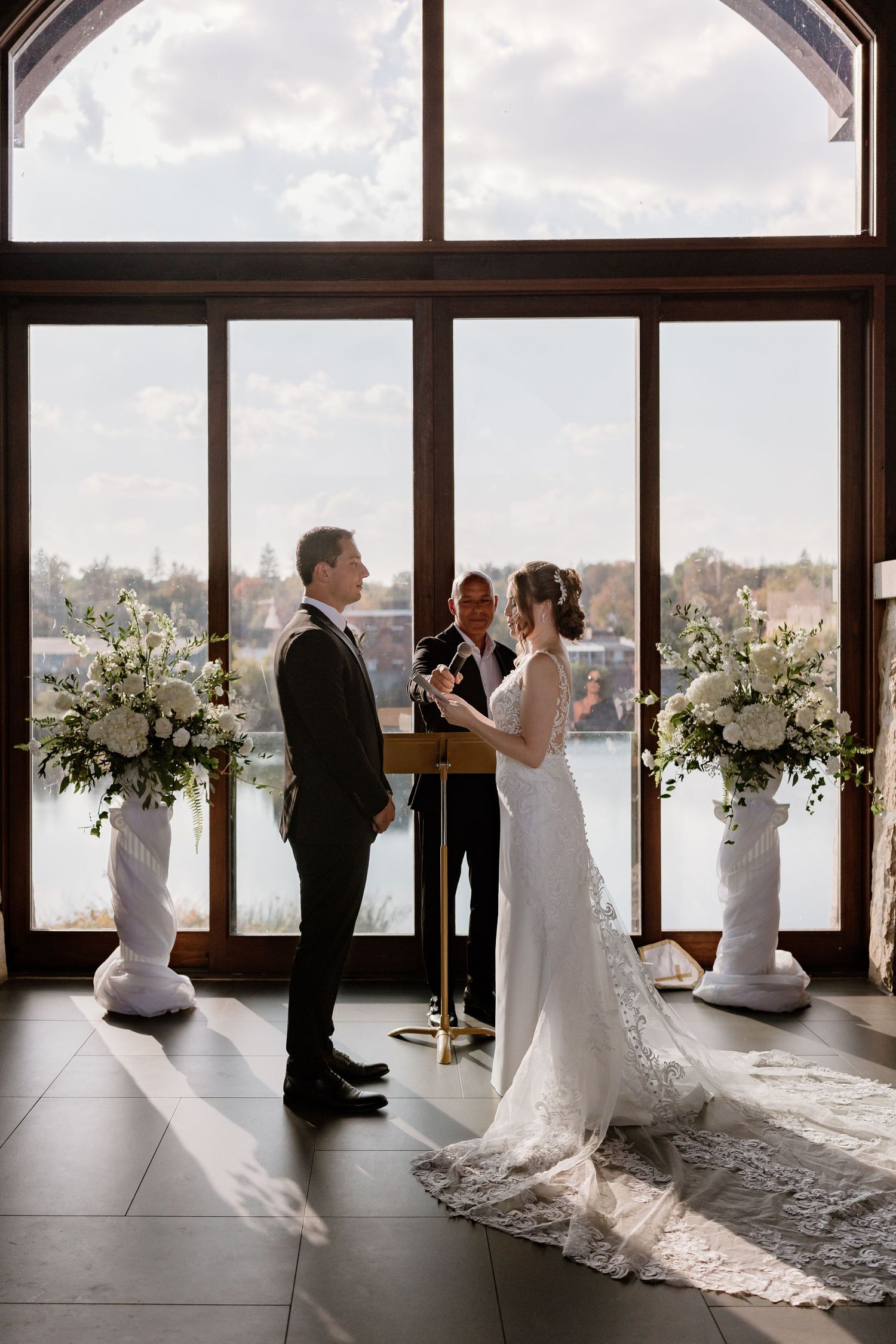 A wedding couple shares an intimate first look moment in front of large windows with white floral arrangements.