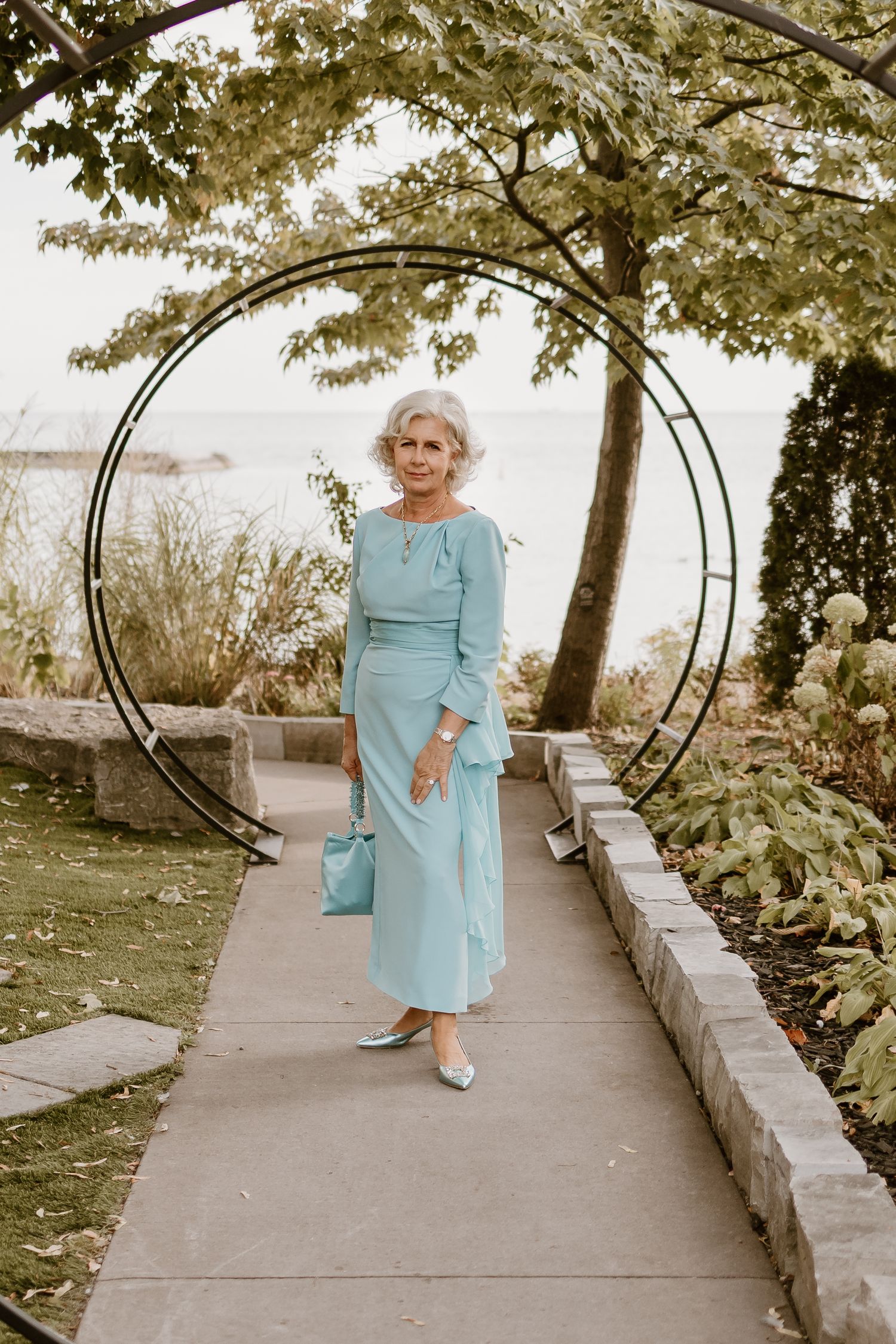 Person in light blue dress posing under garden archway with trees.