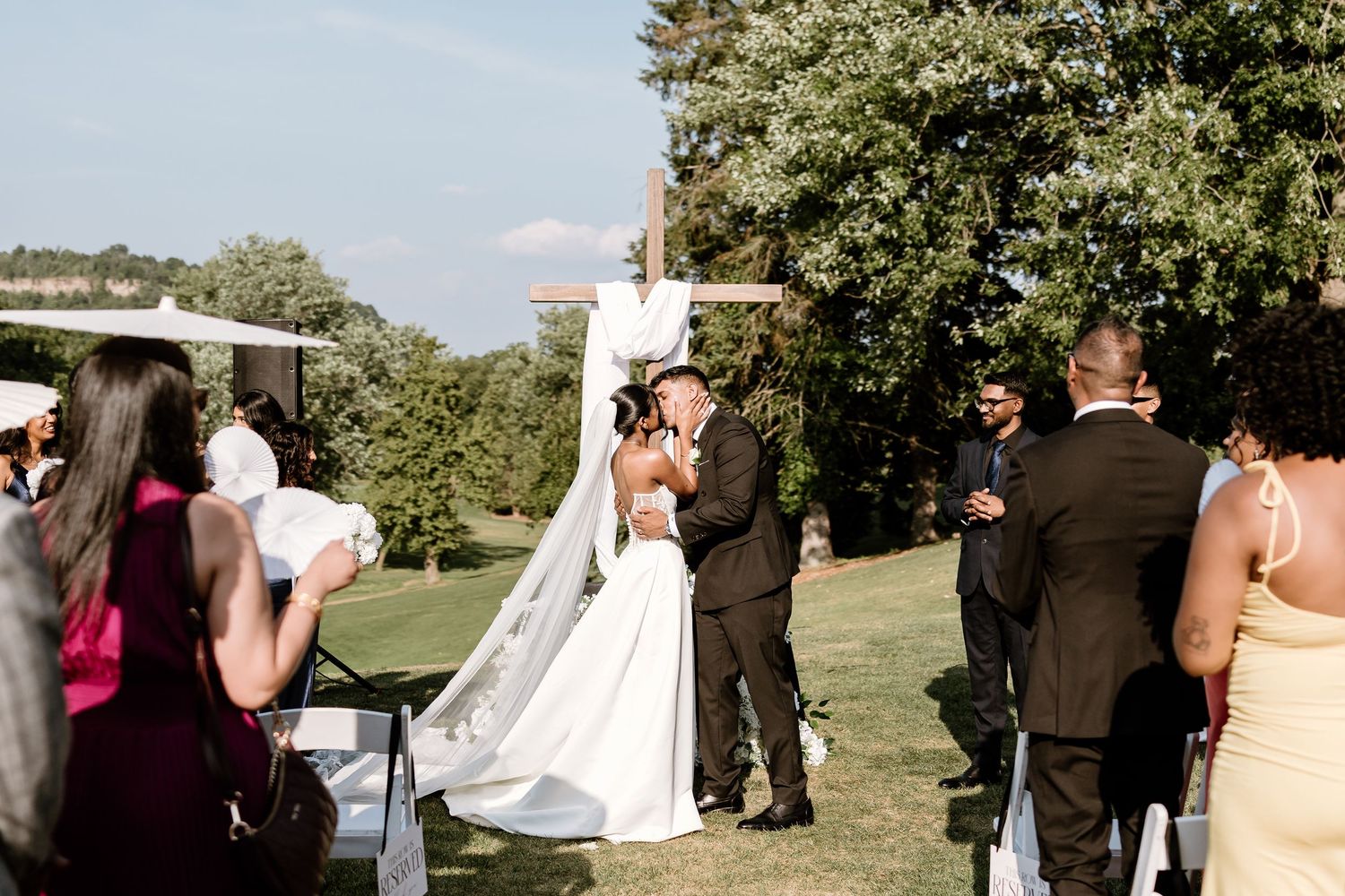 Wedding guests gathered outdoors for ceremony under blue skies and trees.