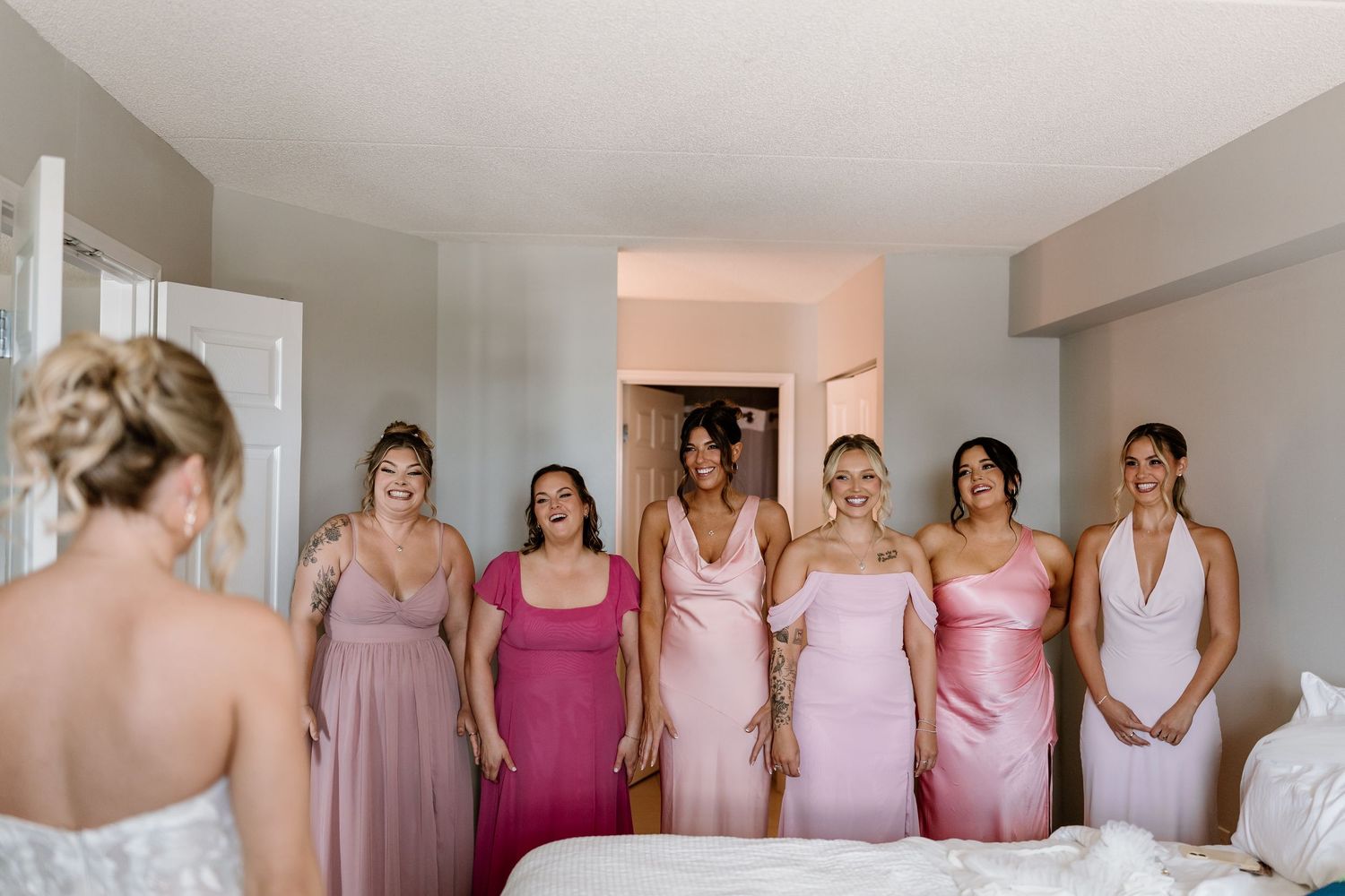 Bridesmaids in pink dresses react with joy seeing the bride in her wedding gown during bridal preparations in a hotel room.