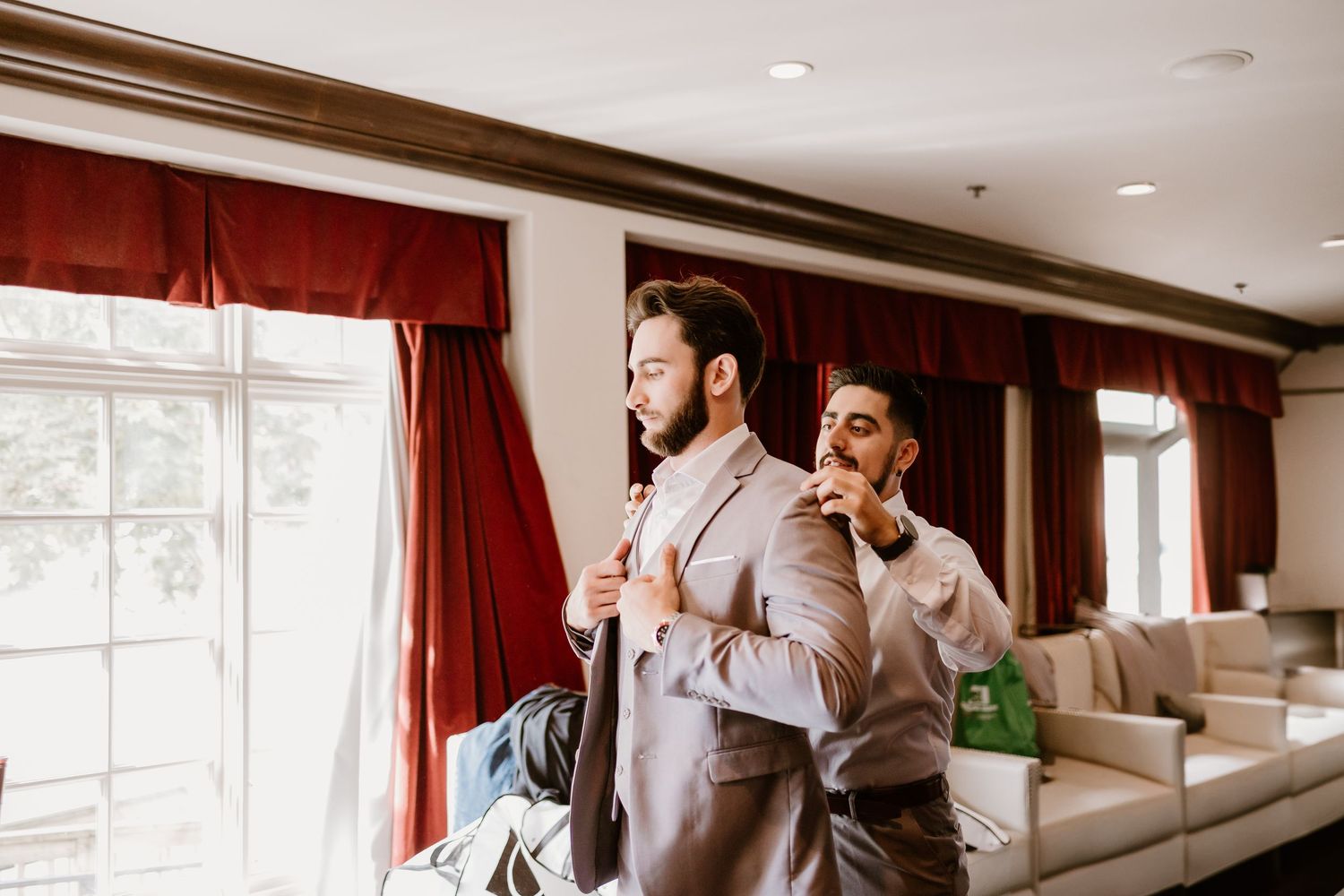 Two people in formal attire adjust their outfits by window with red curtains.