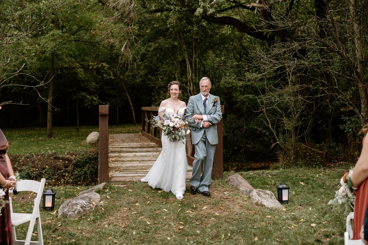 An outdoor wedding ceremony with guests seated on white chairs along a wooded path.
