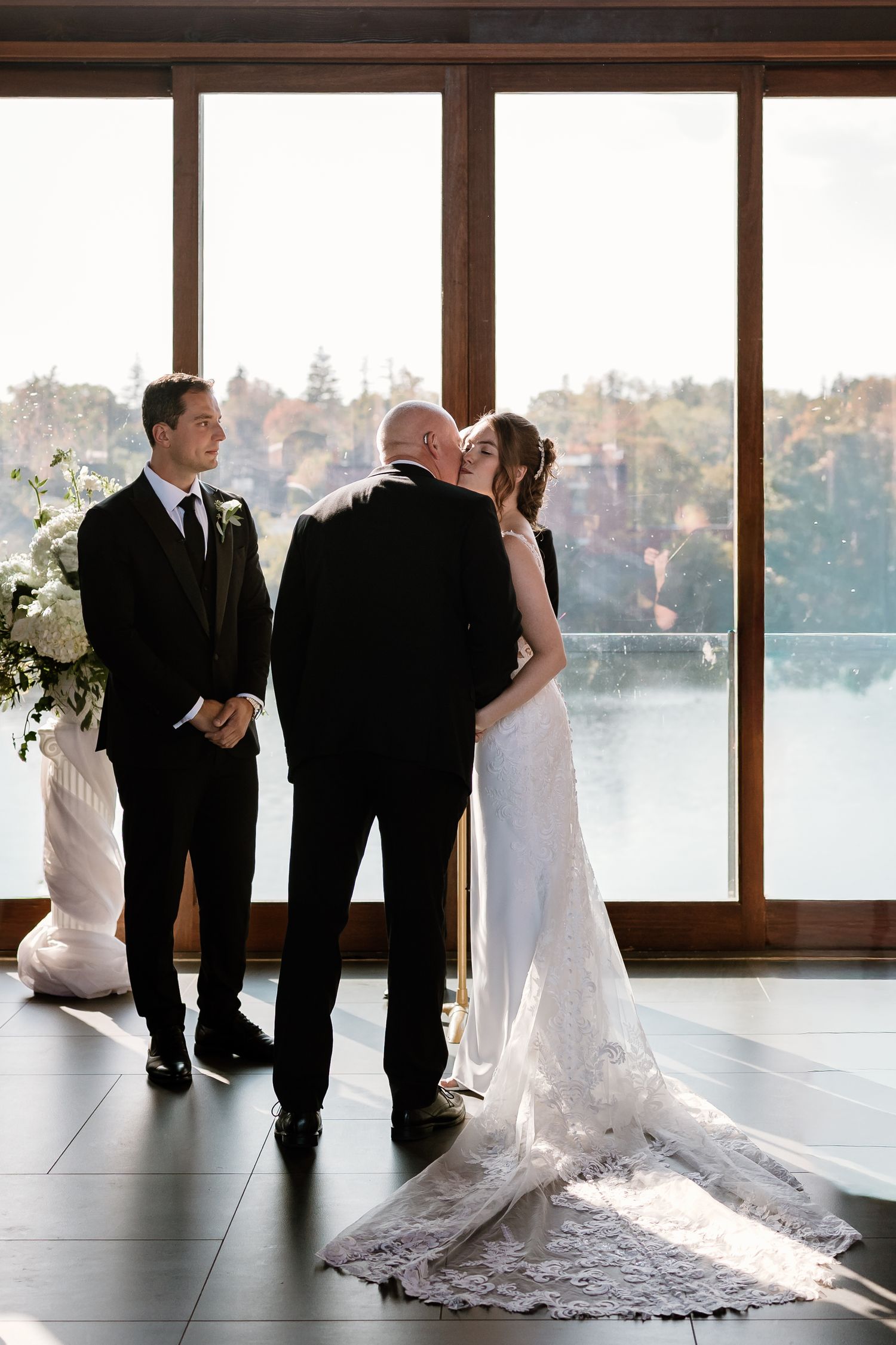 Wedding ceremony in a modern venue with floor-to-ceiling windows overlooking a scenic lake view.