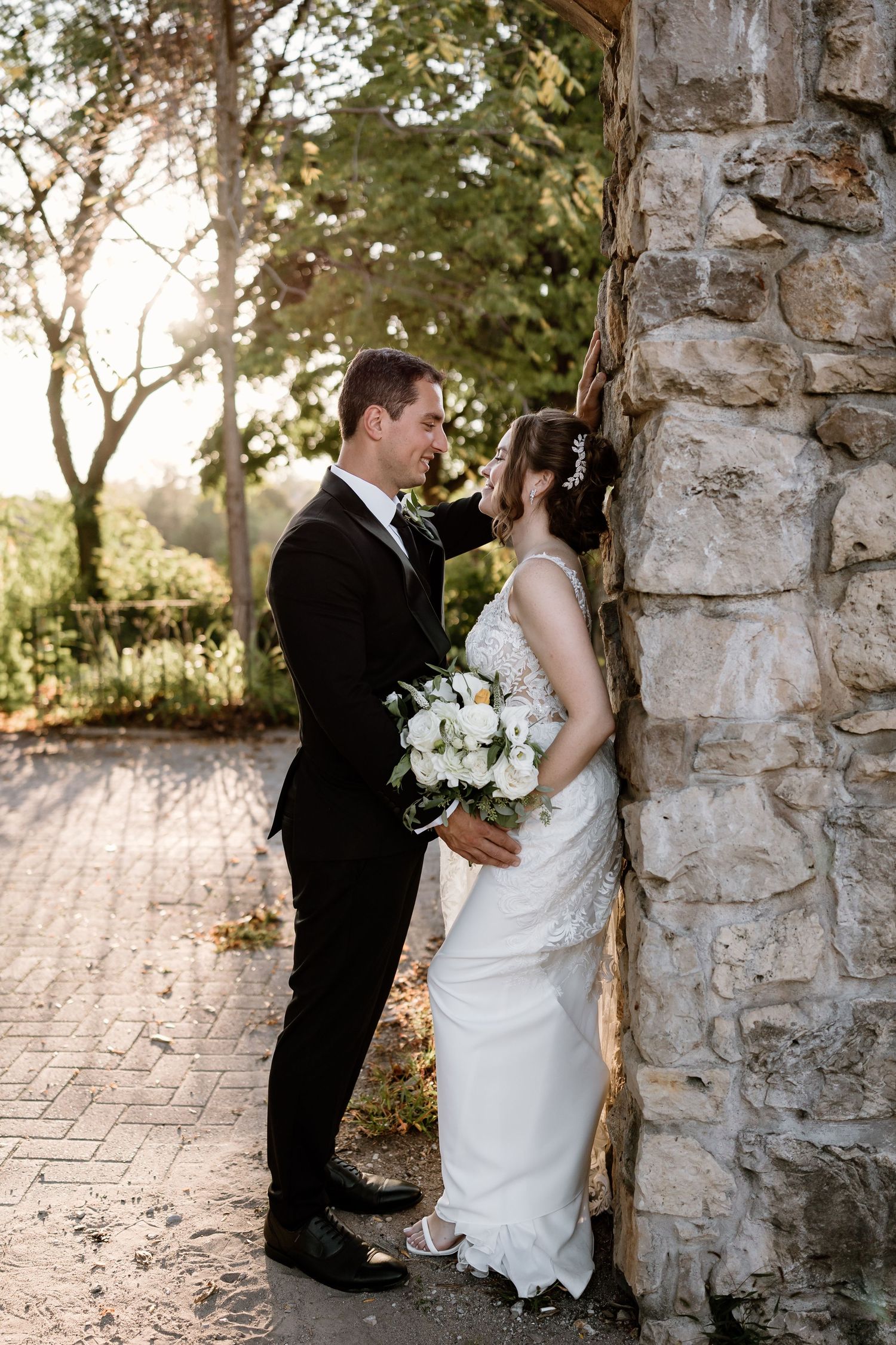 Wedding couple poses against rustic stone wall with white bouquet at sunset.