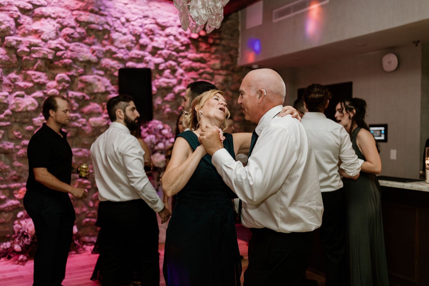 Guests dancing and celebrating at wedding reception with pink ambient lighting.