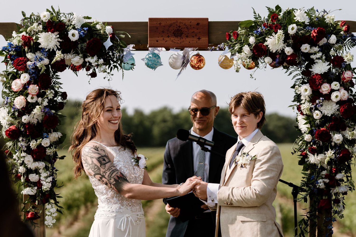 Wedding ceremony under a floral arch with pink and white roses in an outdoor vineyard setting.